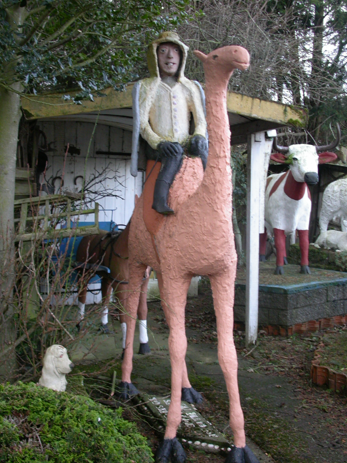 A life size statue of a man - lawrence of arabia - riding a camel, crudely created but impressive. behind him is a statue of a red and white cow with horns, and at the camels feet is a small white dog