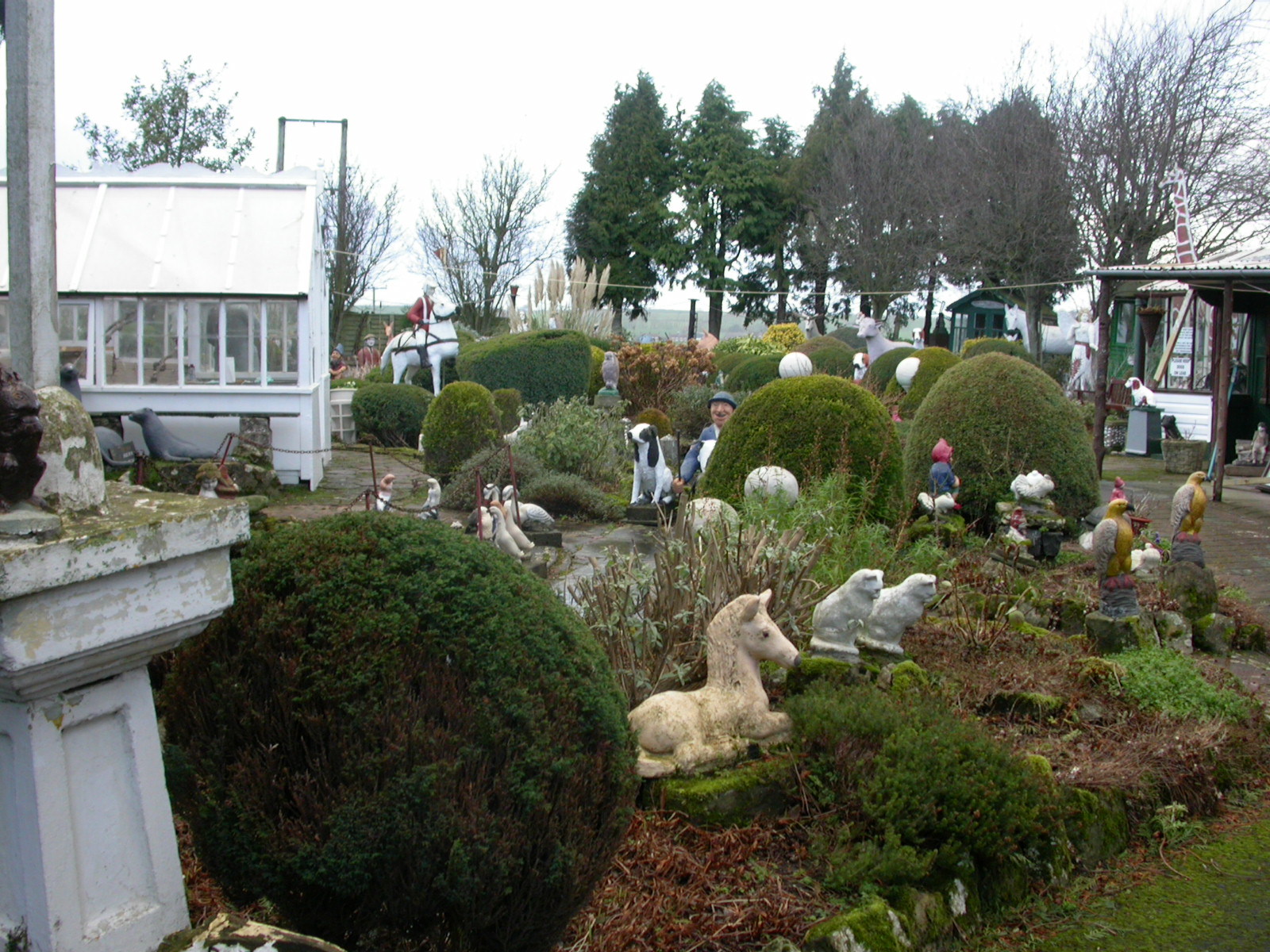 A large garden with immacualte topiaried globe shaped bushes, scattered in between are a large number of small concrete statues of people and animals. There are a few shed like structures too.