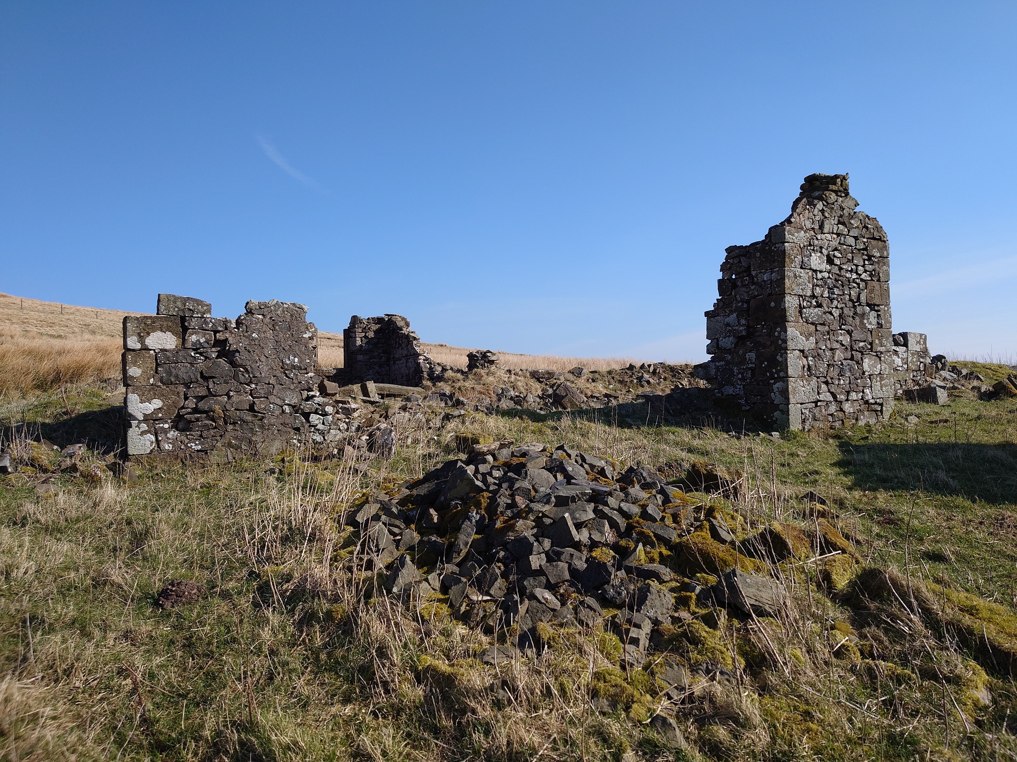 A ruined stone house, very little is standing other than three corners. Stone rubble lies everywhere, grown over with grass.