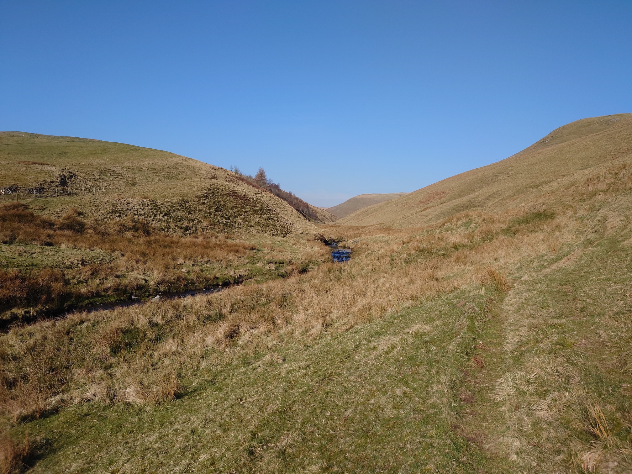 Under a lovely blue sky, a stream burbles through grassy steep sided hills