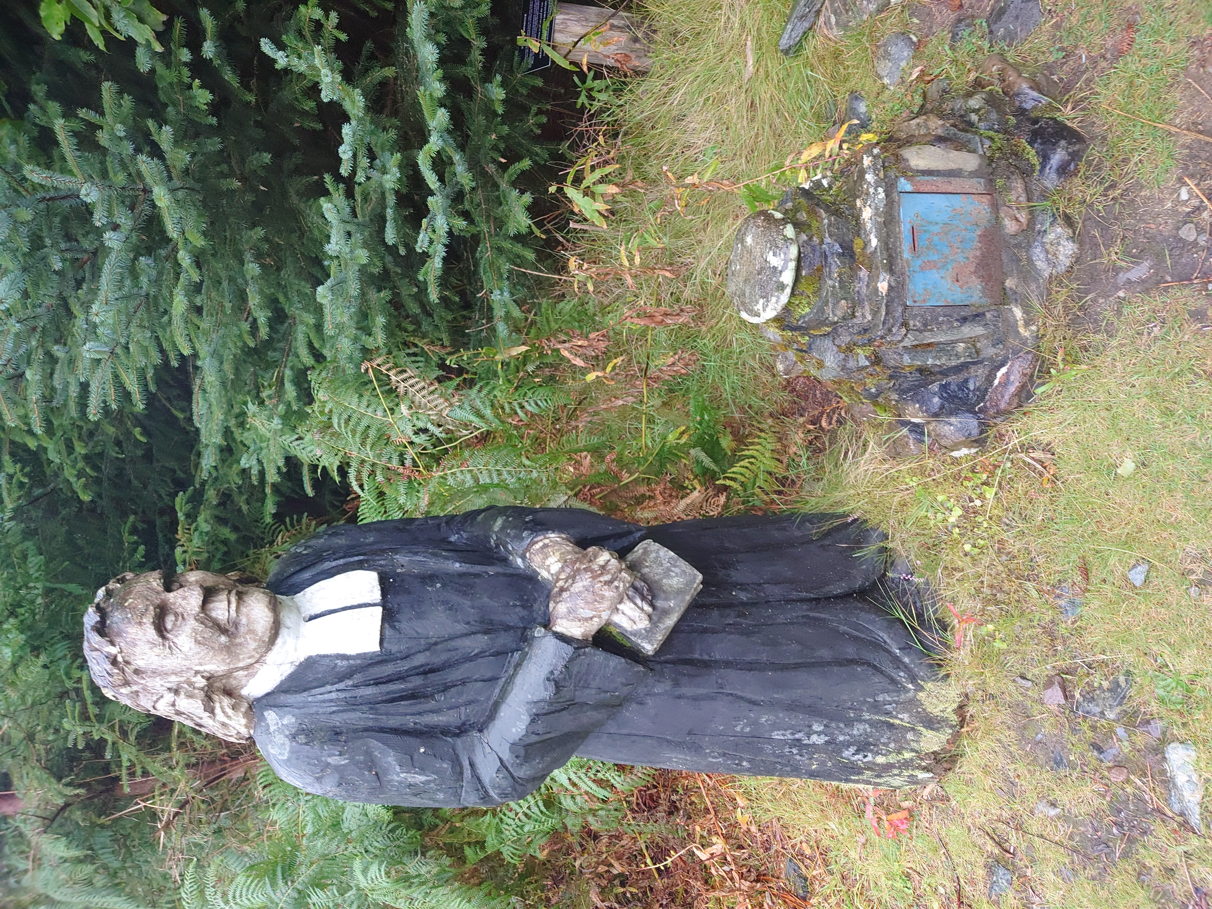 a painted wooden statue of a stern looking man in priest's clothes, clutching a bible, next to a small donations box made of stone.