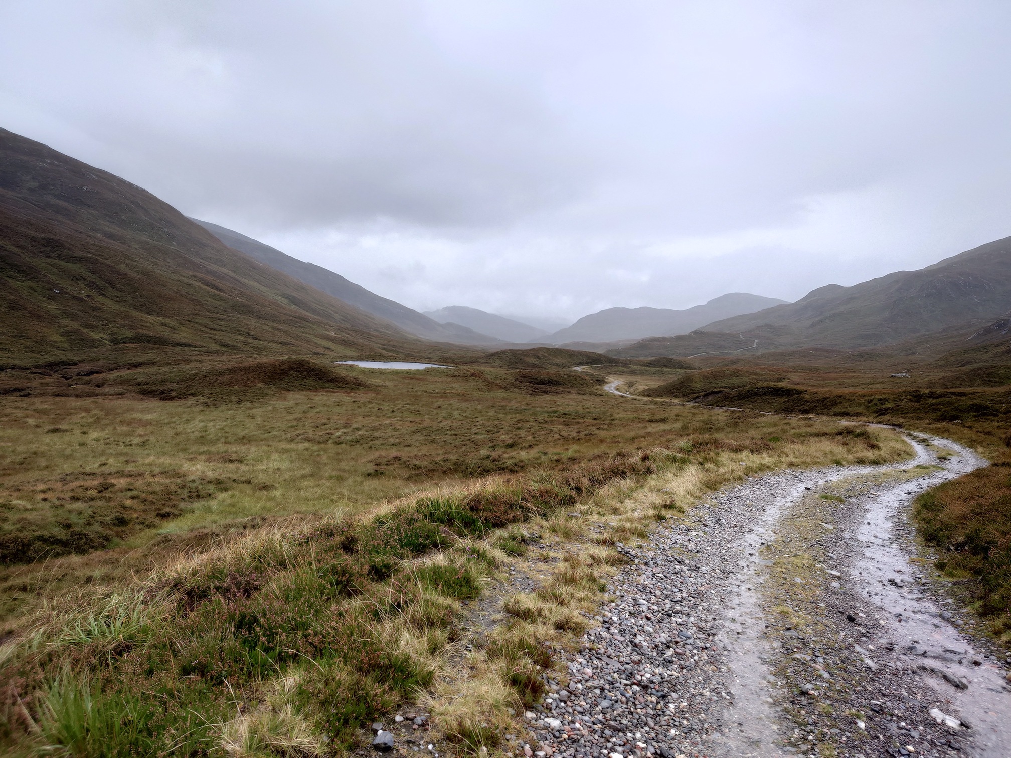 A gravel track winds its way into the far distance over boggy ground in the valley bottom. Either side and in the distance, the hills rise up into the cloud.
