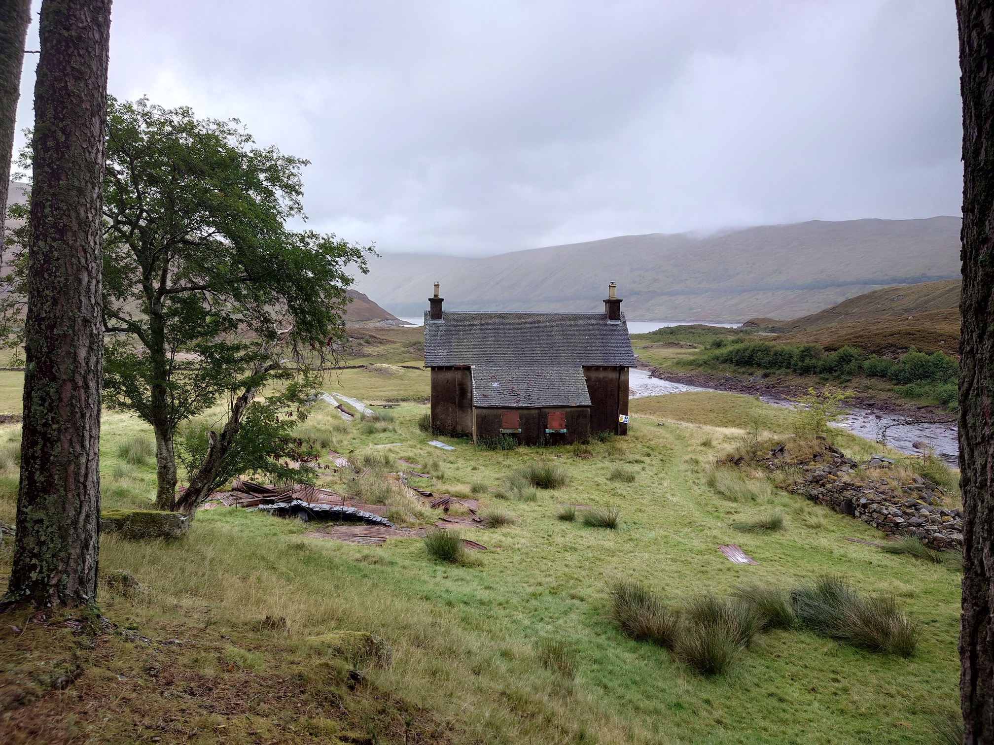 Behind the lodge, a two storey stone house with boarded up windows, looking out from the trees to the loch and hills behind.