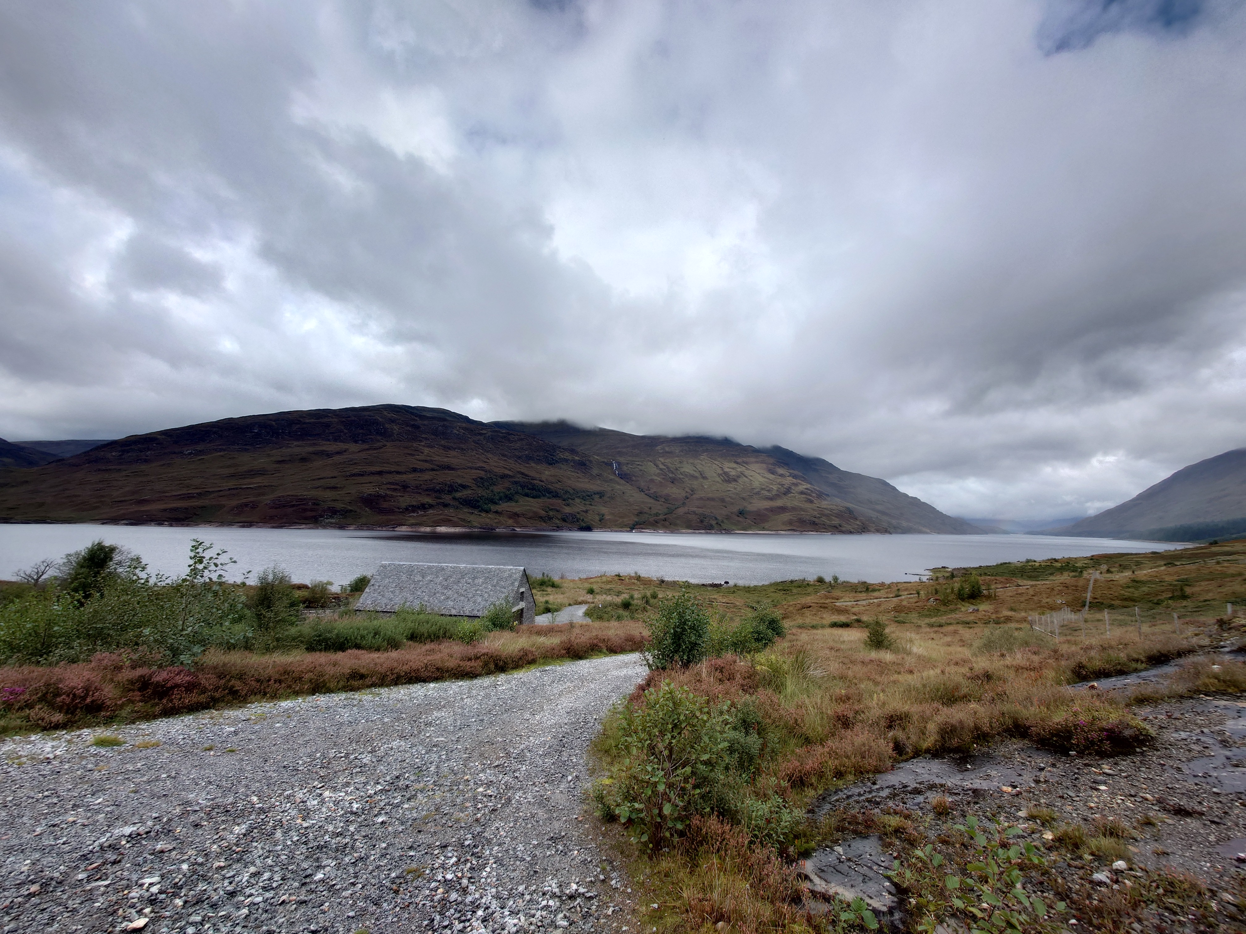 A view of the Loch from a broad gravel track. Below it, the slate roof of a stone building.