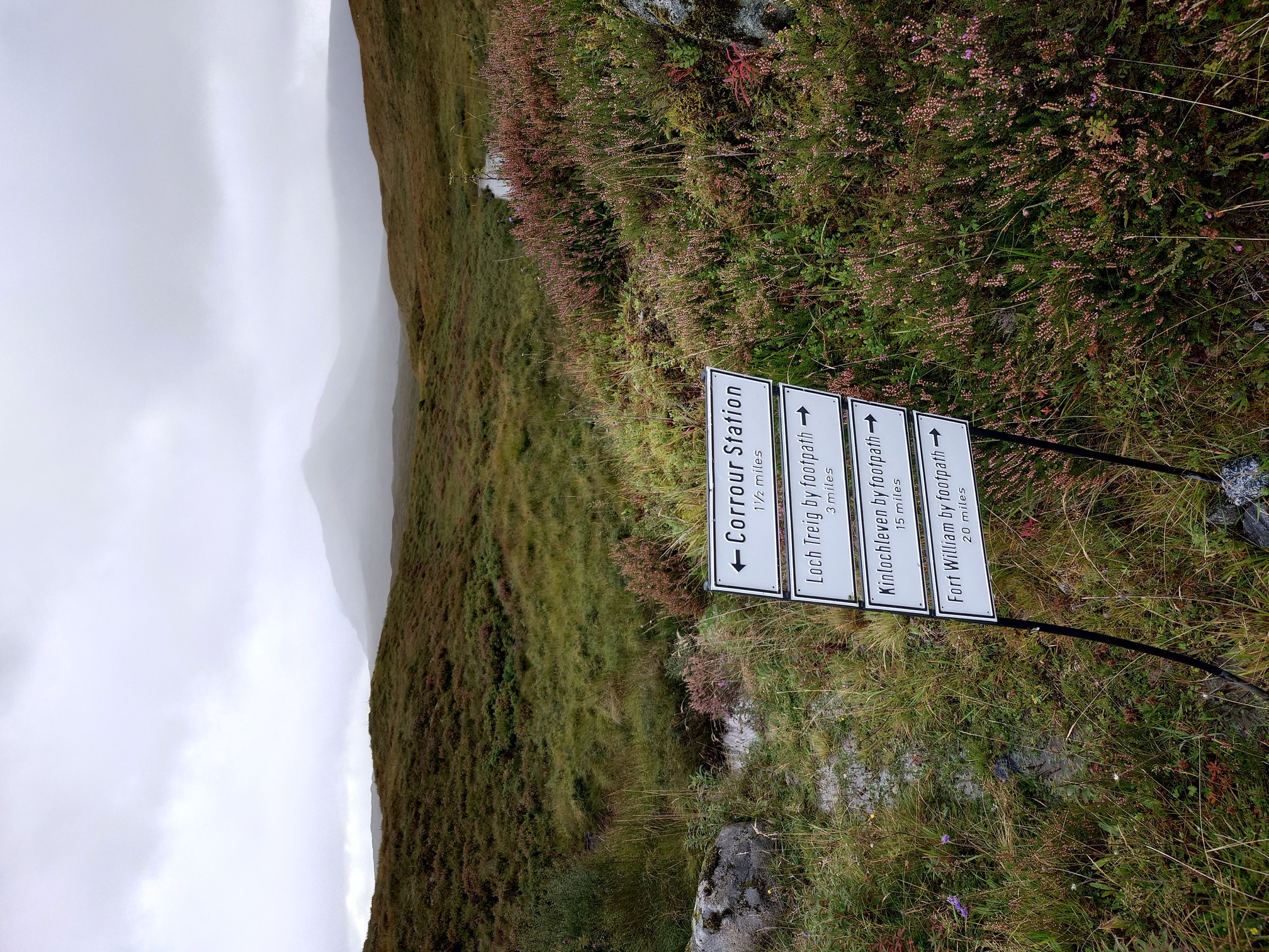 Four white oblong signs with black text, one above the other, on two metal poles in the ground, in the heather and bog. Corrour Station (left) 1 and a half miles; Loch Treig by footpath (right) 3 miles; Kinlochleven by footpath (right) 15 miles, Fort william By Footpath (right) 20 miles