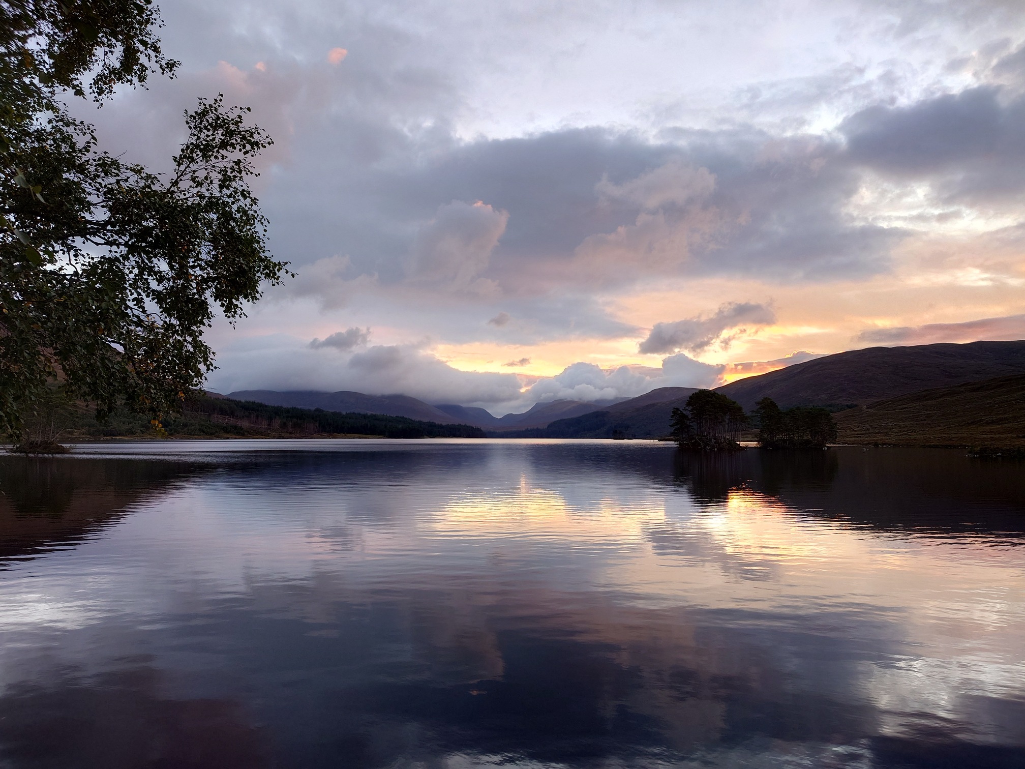 The early dawn sky with hughes of pink and yellow behind the clouds, over the loch with the hillsides in shadow.