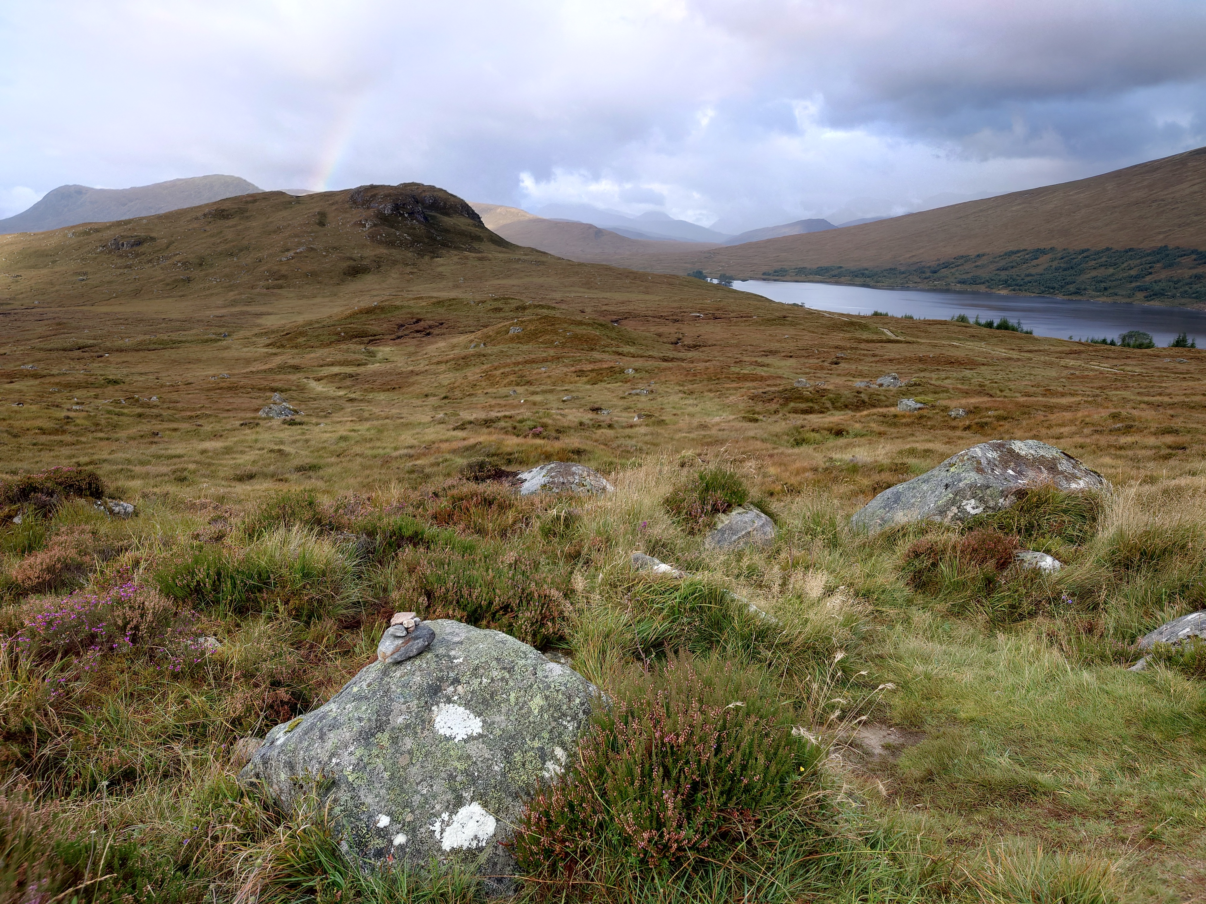 A few boulders in the heather, one has pebbles piled on top and holds the bronze plaque. The land leads to hills and Loch Ossian, and a rainbow is rising in the sky above.