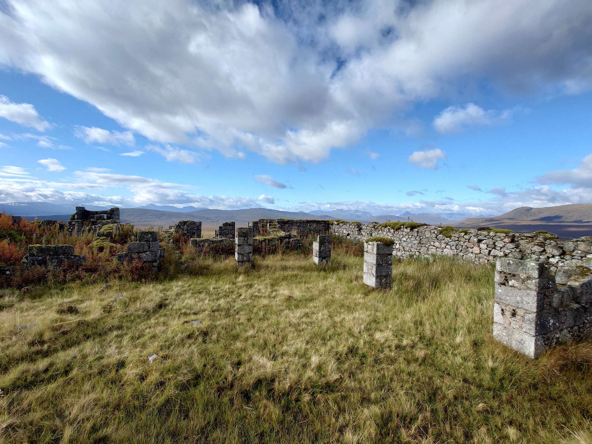 well-made granite stone blocks make up the ruined walls of the lodge. there are a few columns in the centre, but none of the walls or columns are higher than chest height. Mountains can be seen in the far distance. 