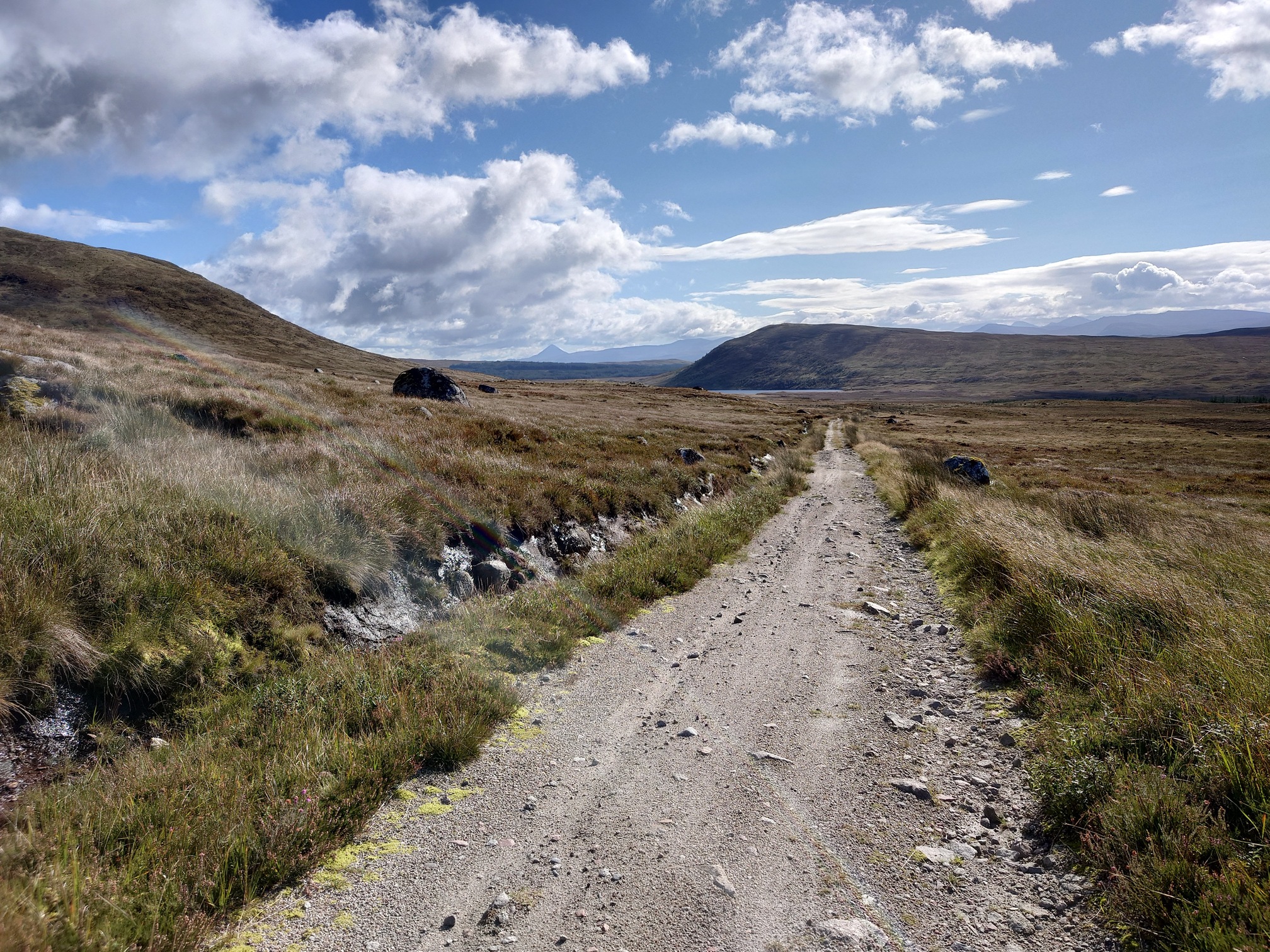 In the lovely sunshine, the road runs straight ahead through towards a small waterbody, behind which is a hill Sron Smeur. Beyond this is a distant ridge, ending in the conical shape of Schiehallion. 