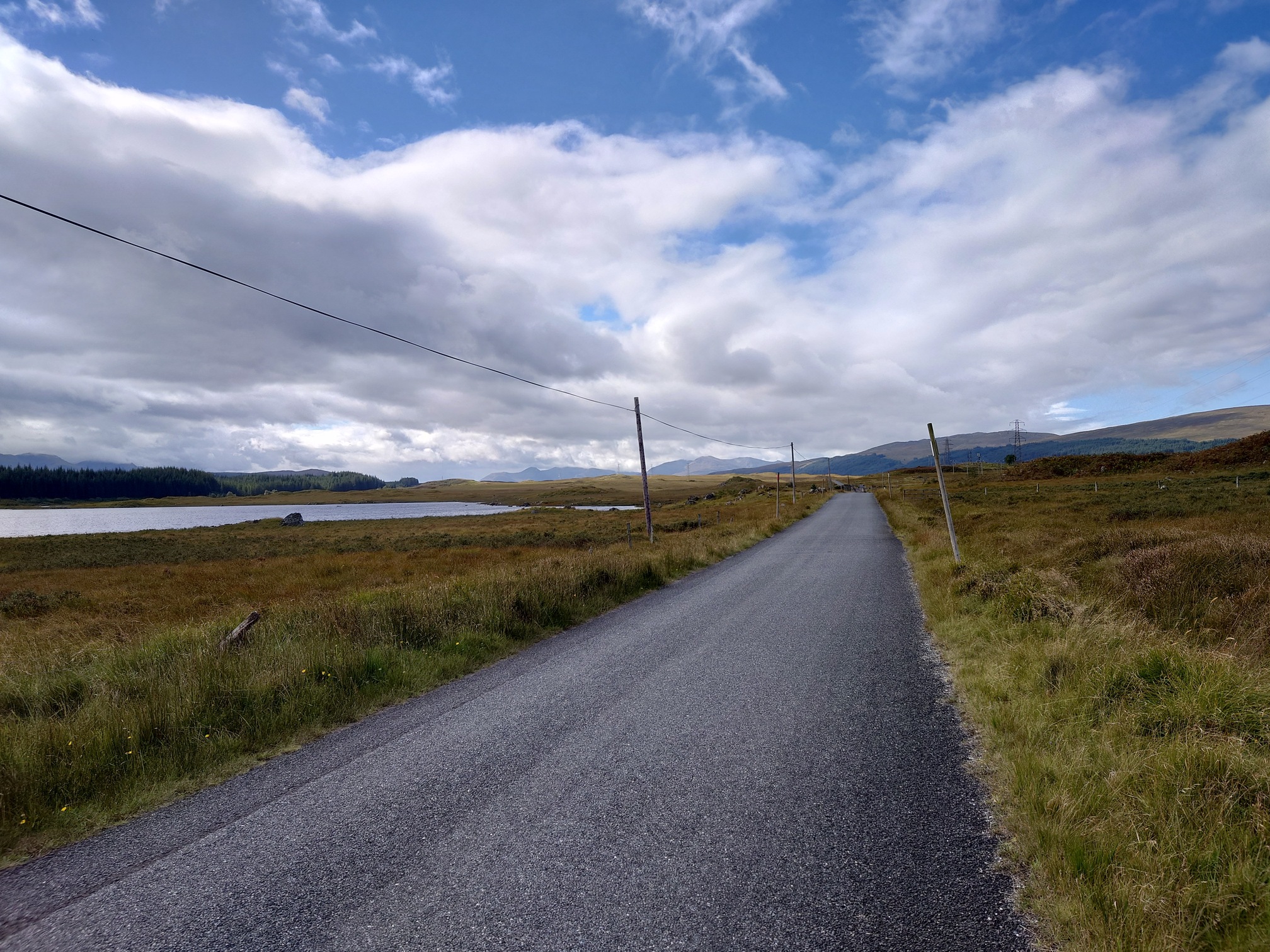A tarmac road runs alongside the loch. Wooden pylons carry a single strand of wire into the distance.