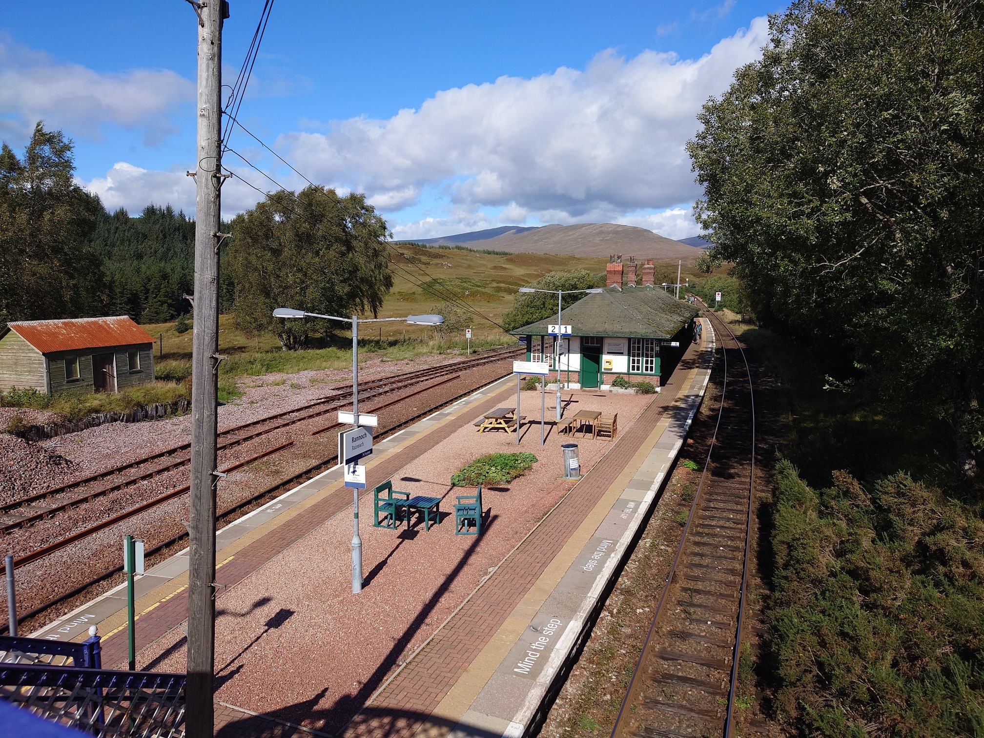 Rannoch Station from the footbridge. An island platform with pink gravel between two tracks, with a single storey, green and white painted station building. There are the usual Scotrail signs and lighting, but nice picnic tables and flowerbeds on the platform. on the far side of the tracks is a wooden hut with a rusty red roof, and there are trees alongside the track but in the distance, of course, are more mountains.