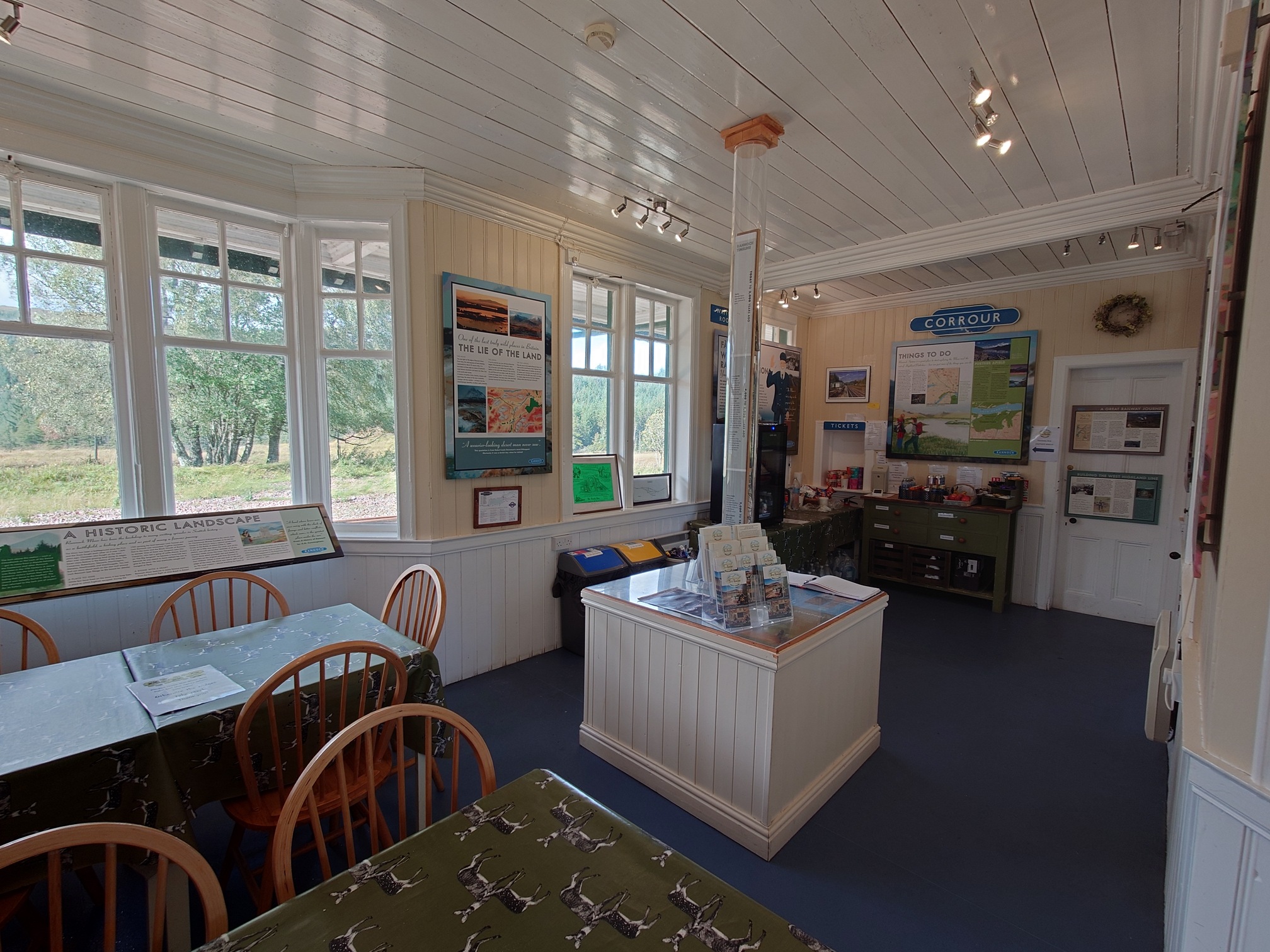 Rannoch Station Honesty Box refreshment room. inside the wooden building there are two tables with shaker-style chairs, a central box with historic information and leaflets on it, and on the far side of the whitewashed room, a rable full of drinks and snacks, a kettle and a fridge. The walls are covered in informative interpretation boards, all about the history of the station and the line, and the ecology of Rannoch Moor.