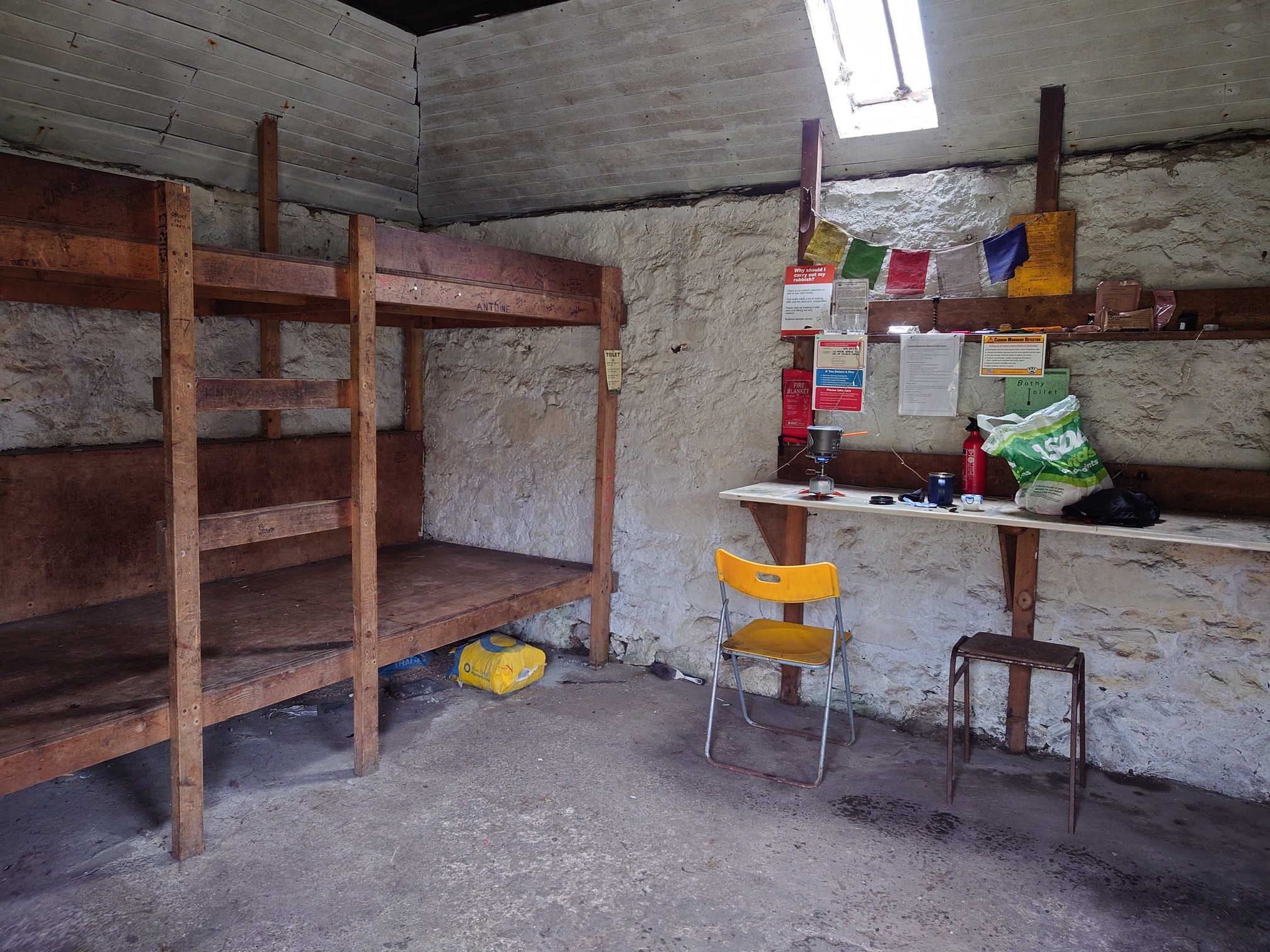 Inside the bothy, the walls are whitewashed and light comes from a single pane of glass in the roof. To the right, sleeping platforms - simple wooden bunks - are covered in grafitti, while on the right notices and signs hang above a white shelf. On the shelf I have my jetboil stove cooking away, and a yellow chair and a stool stand by.