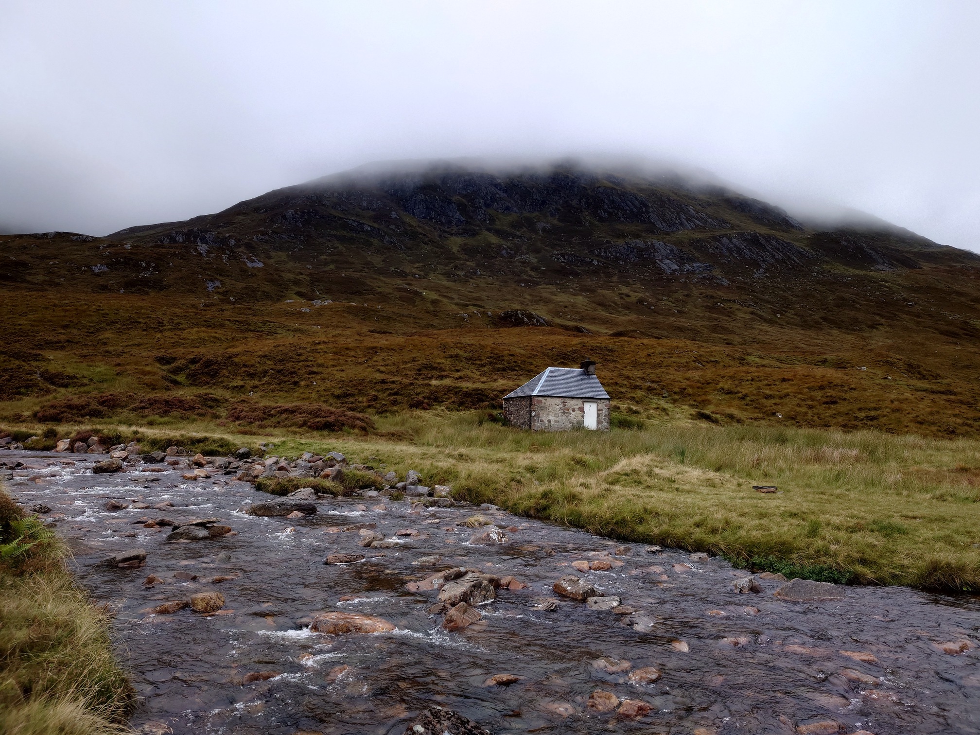 A small, single storey building with only a door, sits on the other side of a wide, shallow, rocky burn. Behind it, a hill brown with heather rises up into the clouds.