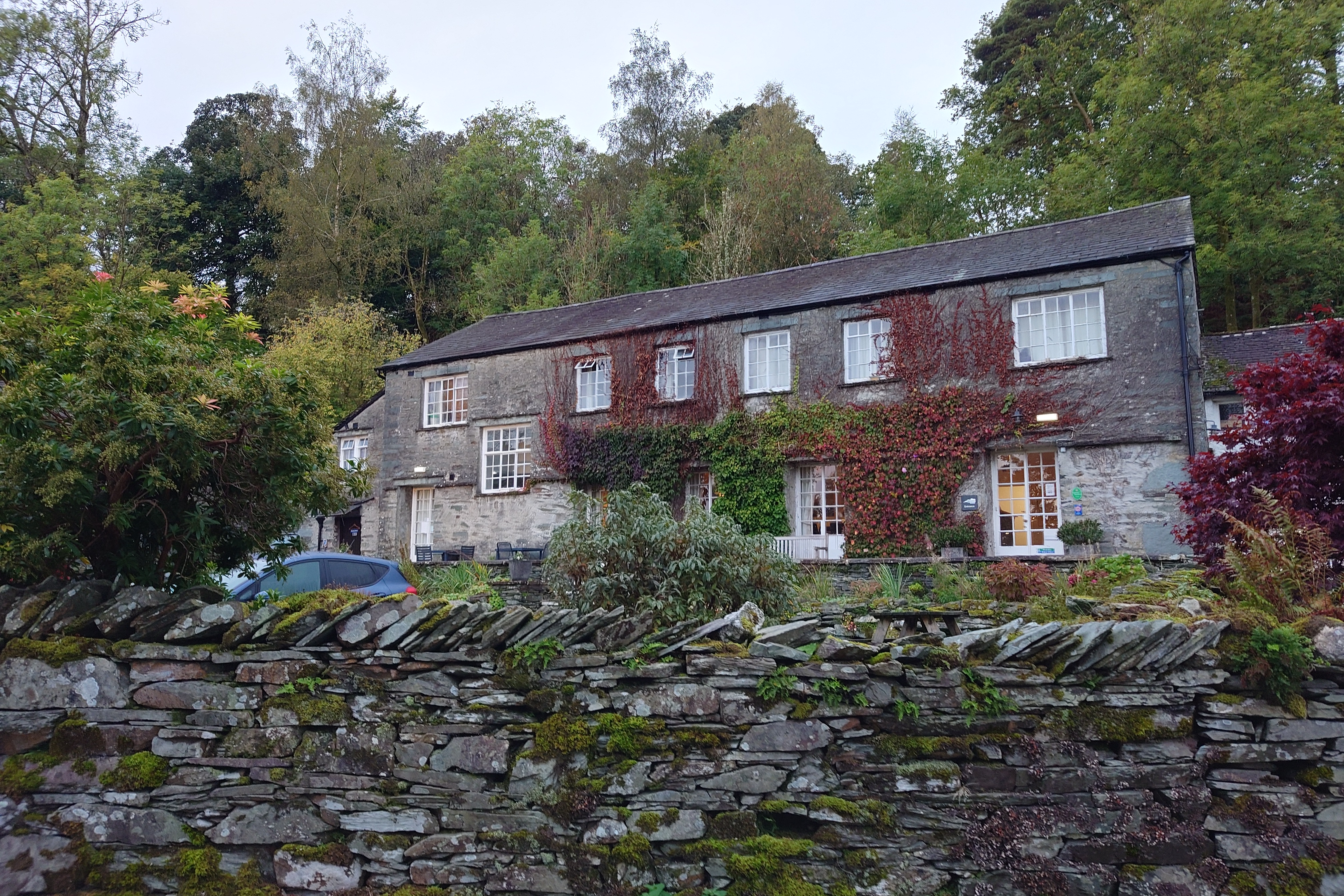 A row of stone buildings that has been knocked into one, with two floors and 6 windows on teh top floor, and a mix of windows an doors on the ground floor. It is covered in Ivy and surrounded by trees.