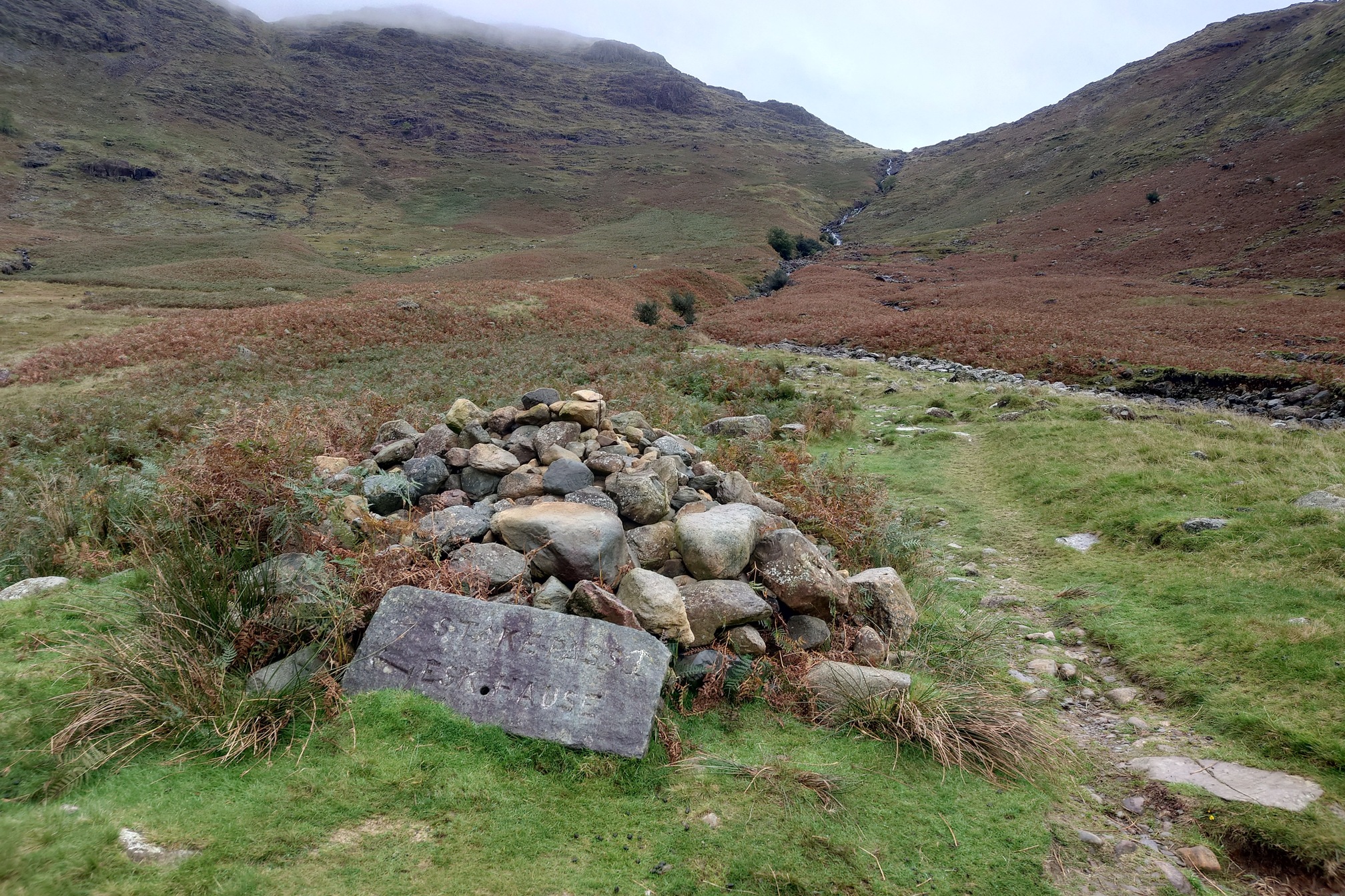 A pile of rocks marks the footpath up out of Langdale to the Stake Pass. The way follows a cascading stream coming down the steep slopes, and the tops are cloaked in cloud.
