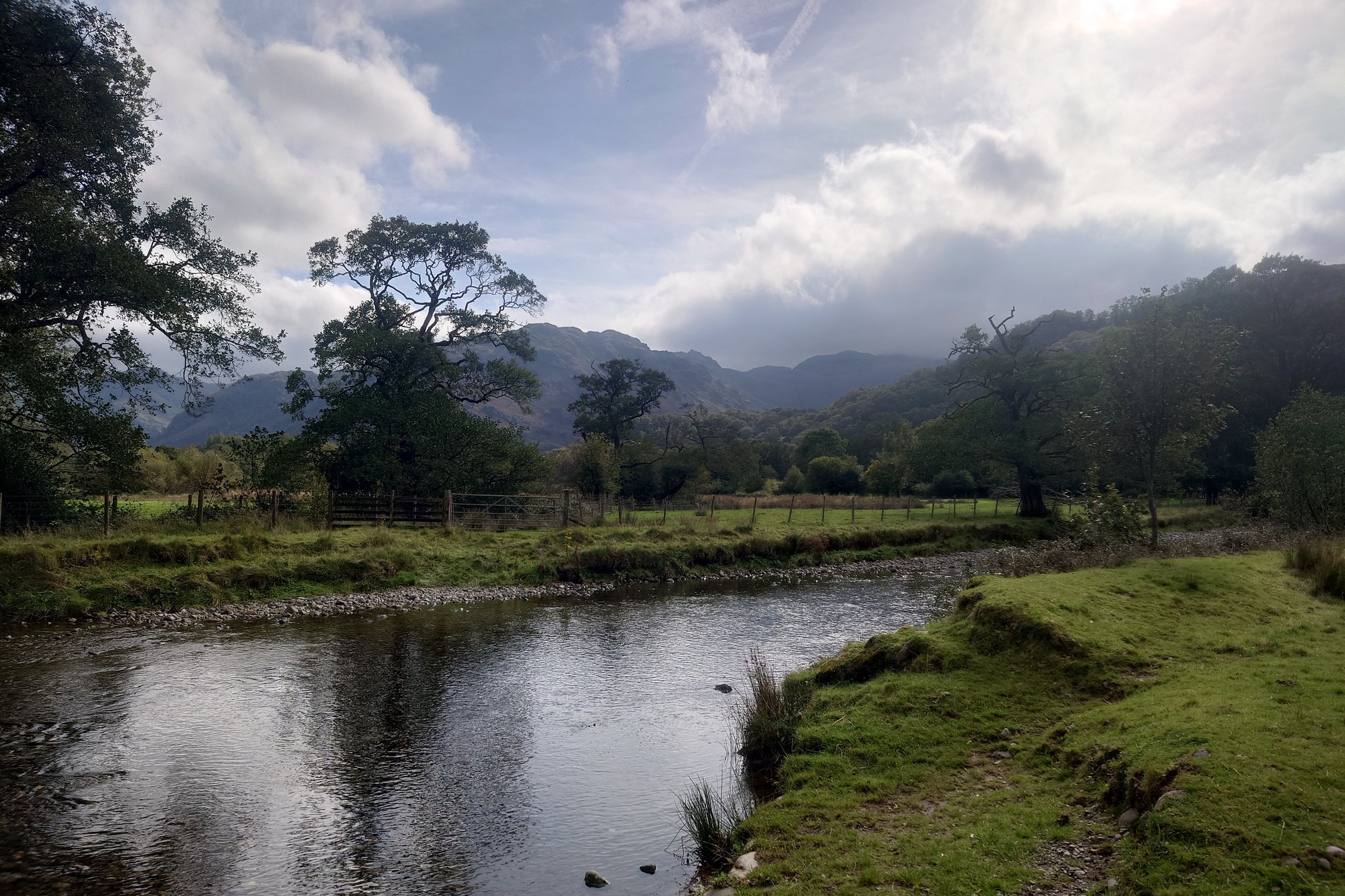 The Derwent River before it reaches Derwent Water. It flows between grassy banks with broadleaf trees, the hills rising in the distance are cloaked in fluffy clouds as the sun tries to break through