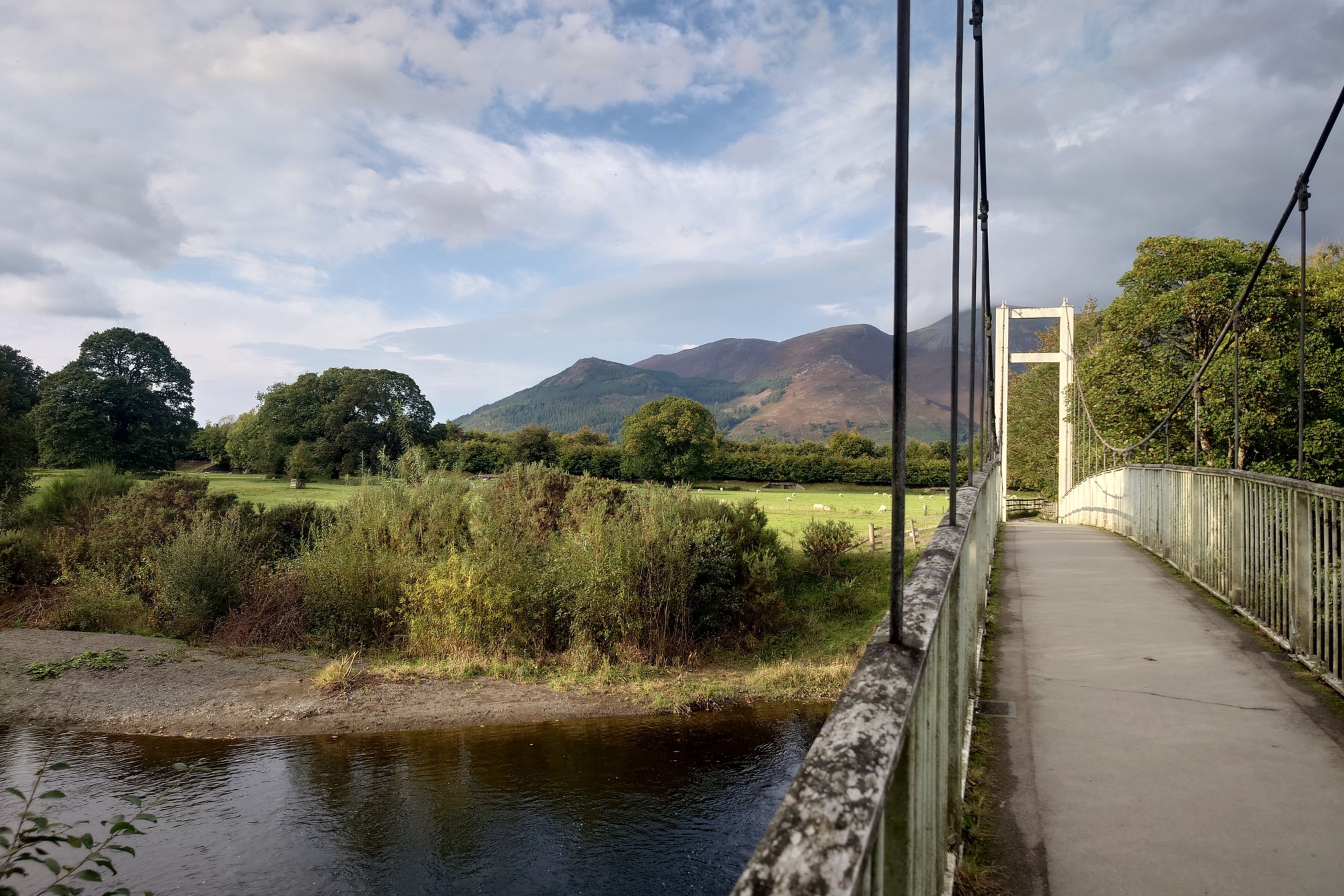 A footbridge over the river. Trees border the river bank seperating it from a grassy field, and of course the hills beyond.