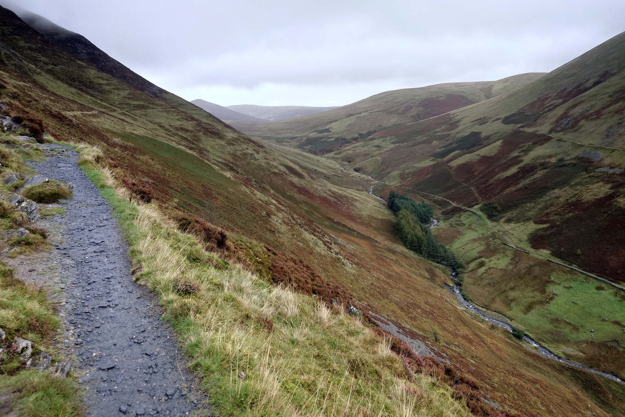 The path heads along the side of a V shaped valley, far above the stream below. The hillsides are brown and yellow with grass and bracken