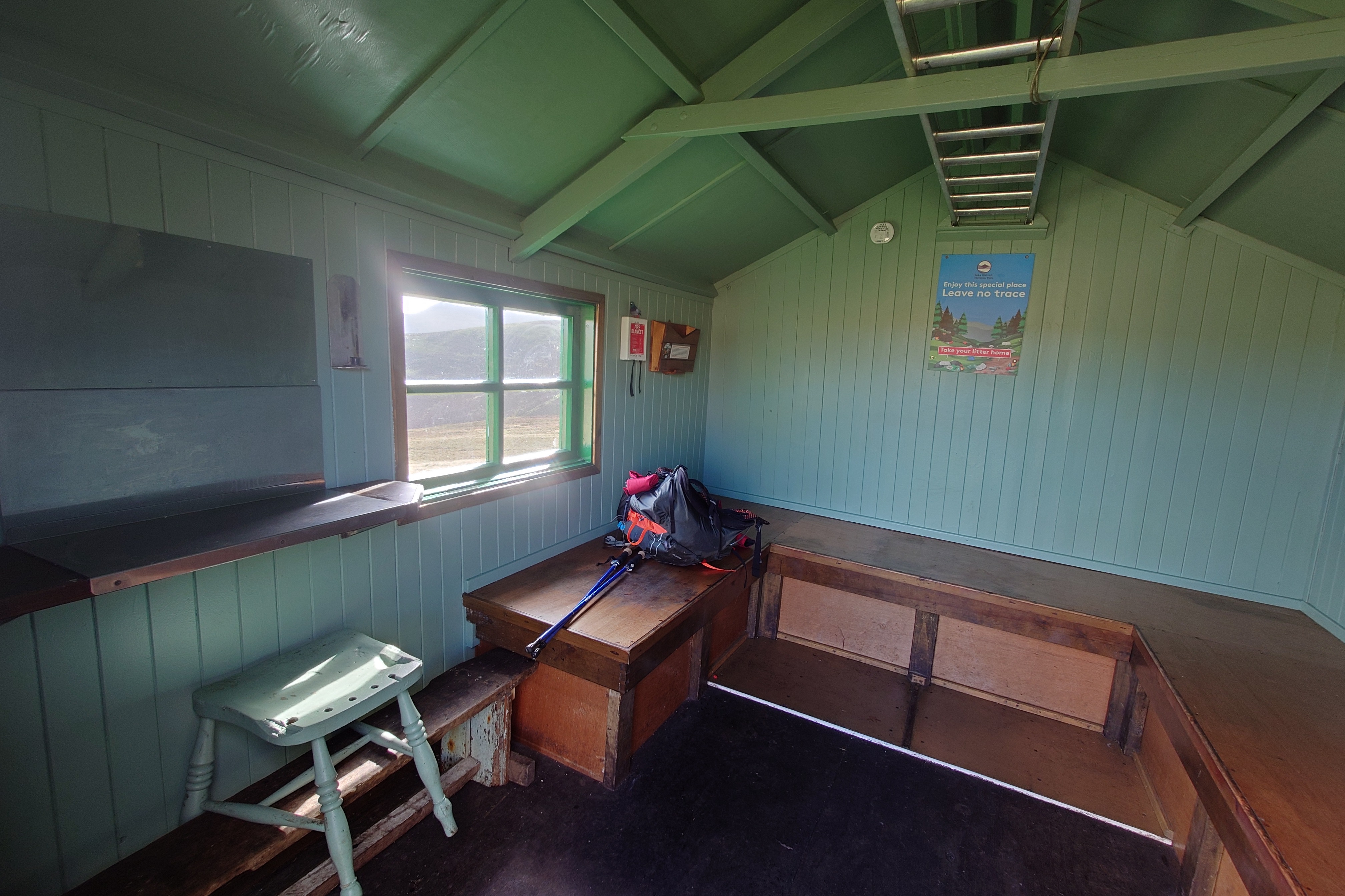 Inside, the wooden hut, the walls and rafters painted pale green while wooden sleeping benches run along the far wall and part of the side walls. A wooden chair sits at a shelf and there is a window providing ample light