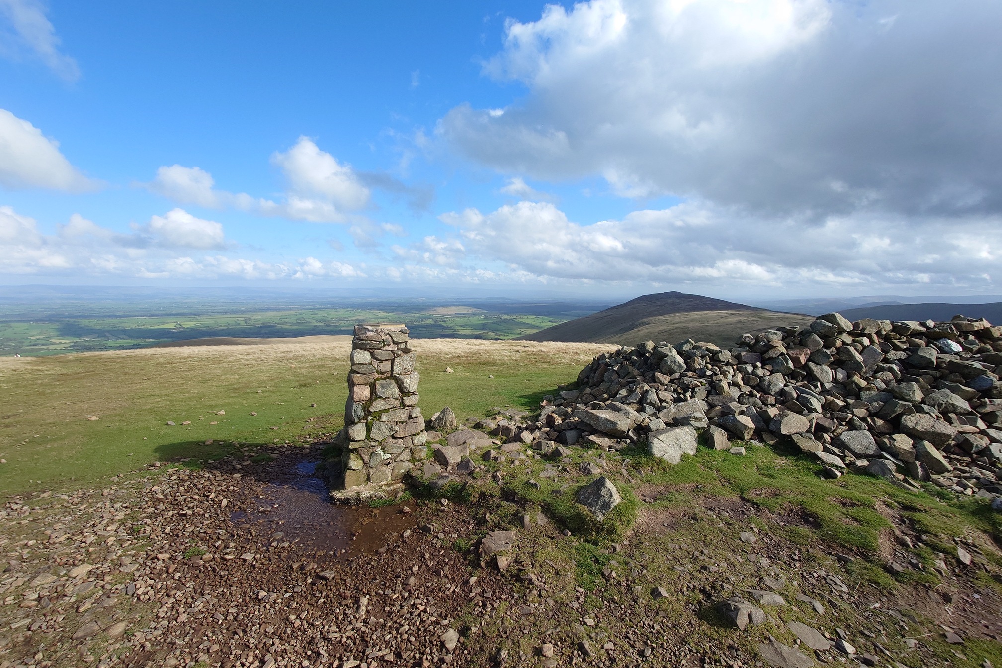 A stone trig point, a pillar about 1m tall, stands next to the pile of stone that is the ancient cairn on the top of high fell.
