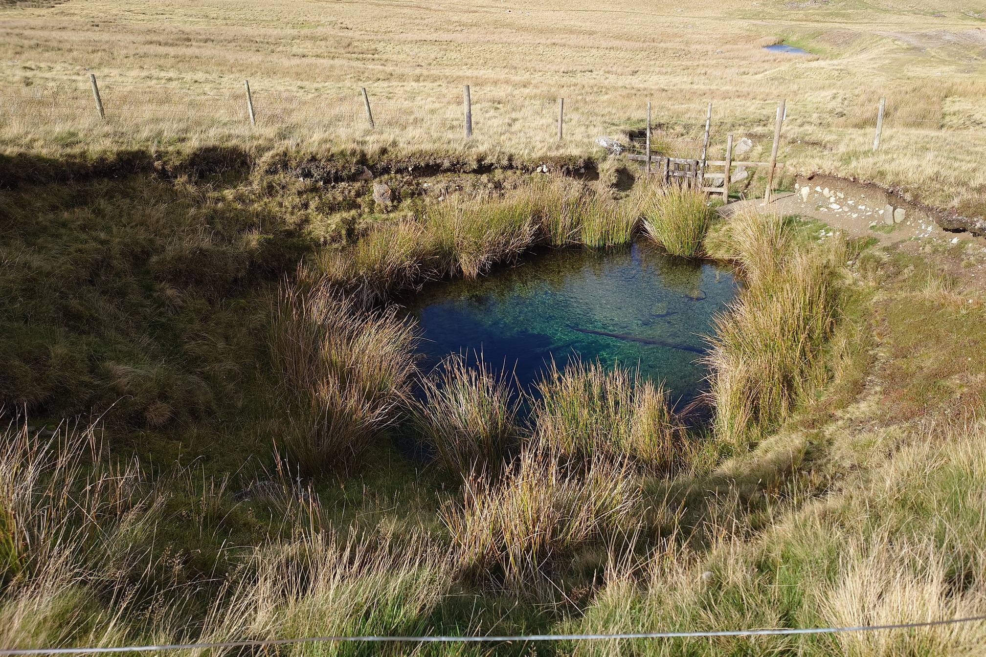 A circular depression in the ground, fenced off, is filled with crystal clear blue water, an old mine shaft