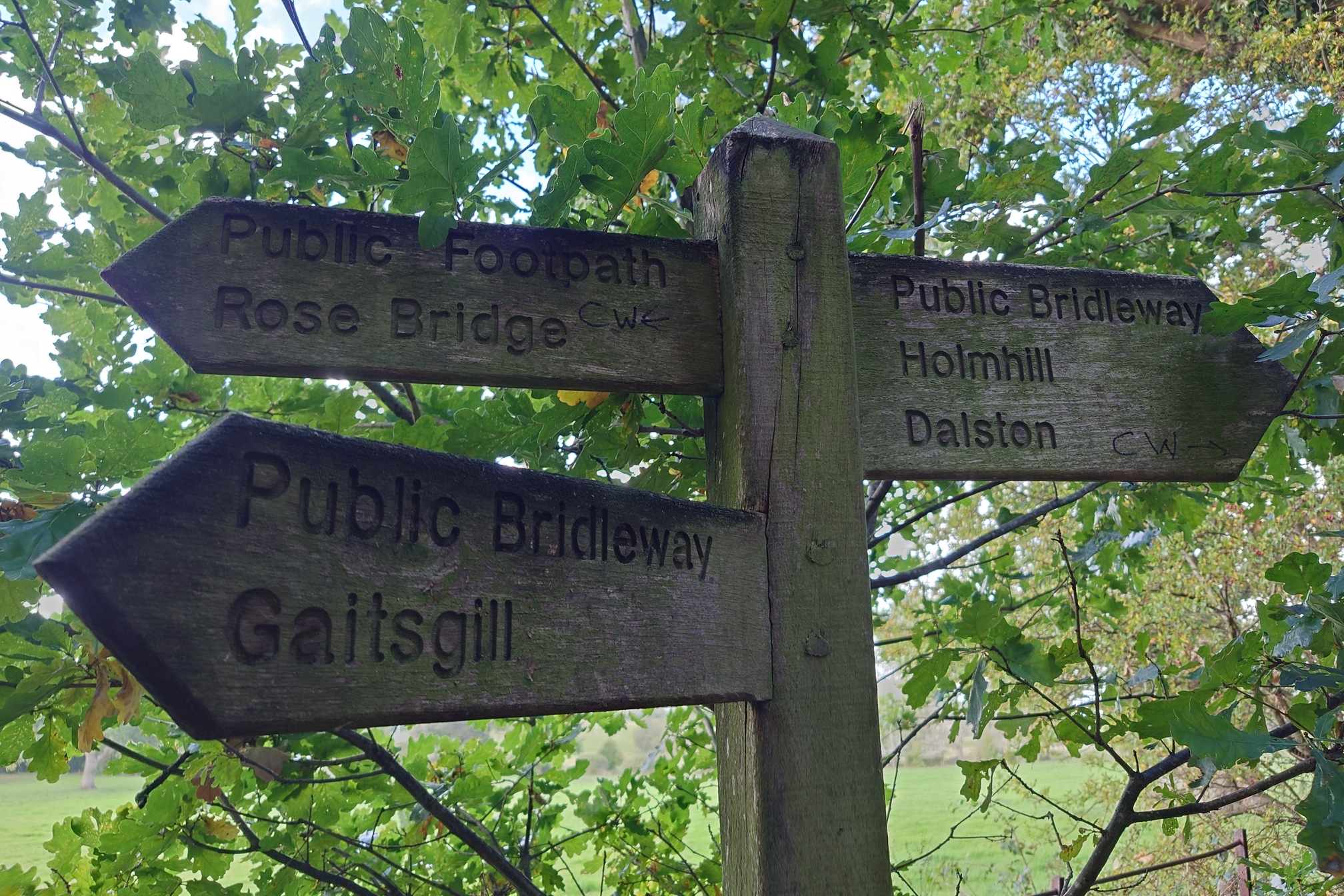 Wooden directional sign pointing in three directions for Public Footpaths and Bridleways, to Rose Bridge, Holmhill, Dalston and Gaitsgill. On two of them the letter CW and an arrow have been scrawled.