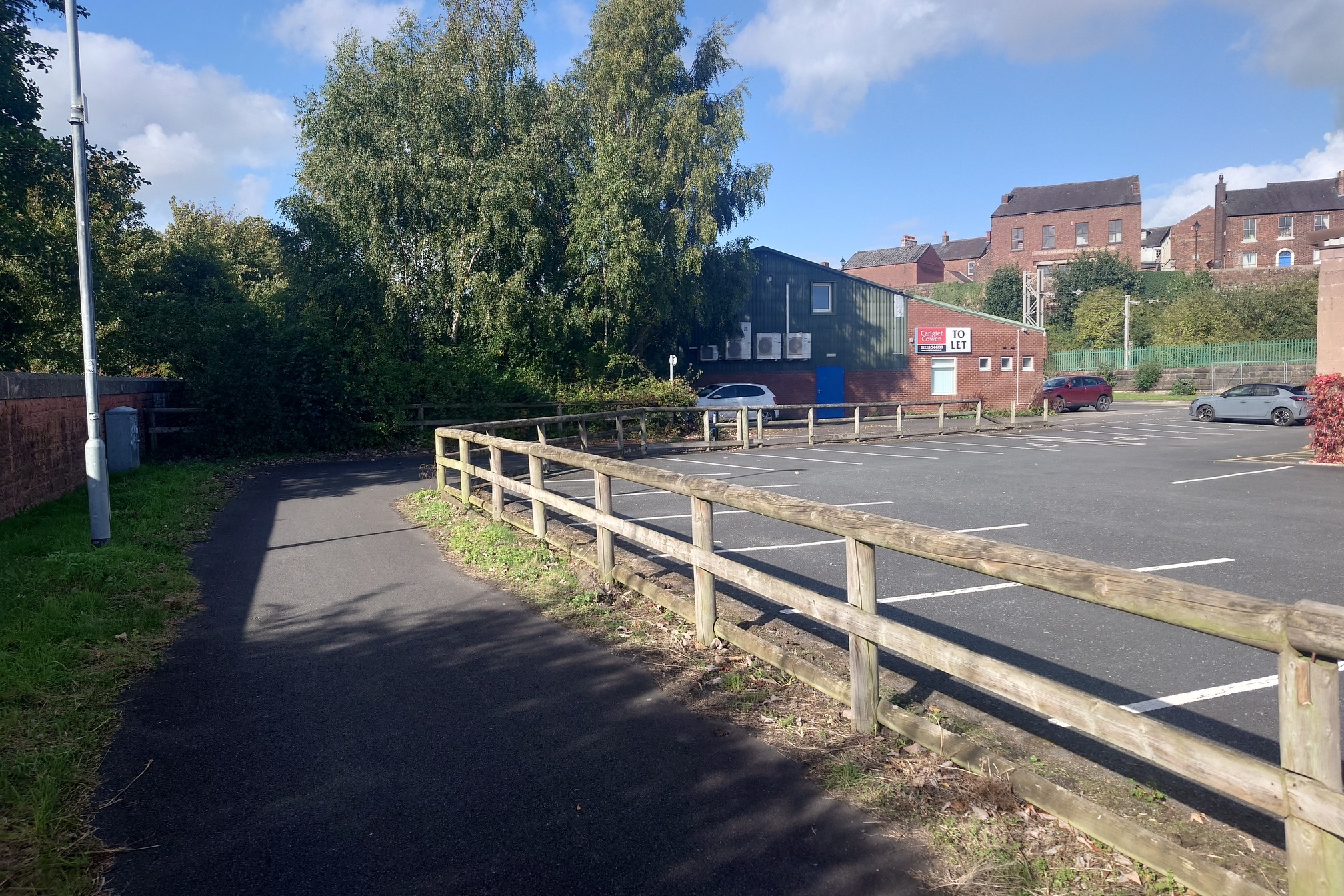 A wooden fence seperates the path from a near empty car park in an industrial estate