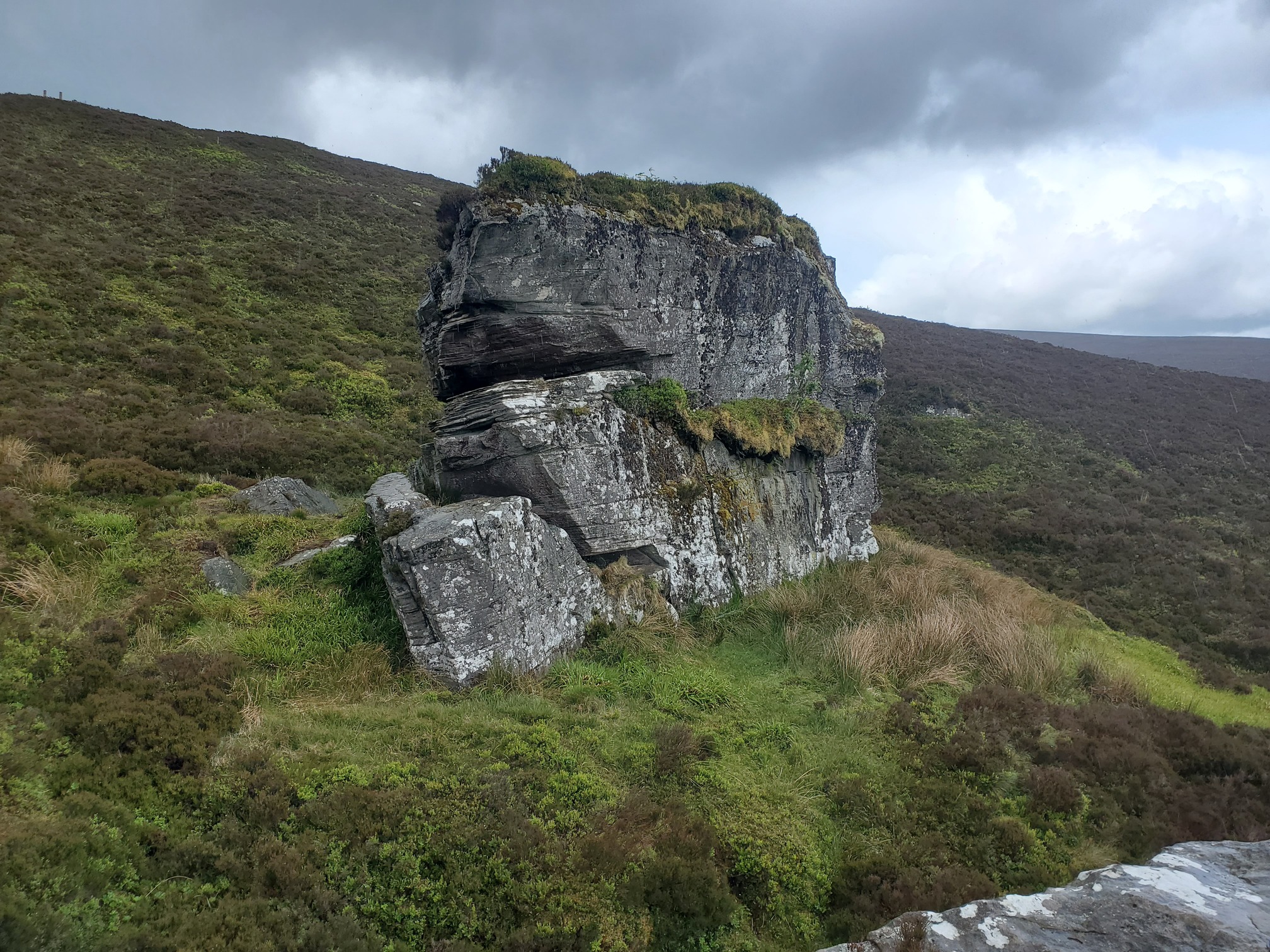 A huge block of sandstone with horizontal clefts. It sits in teh grass on a heathery slope, and has a thick layer of vegetation on top and on a shelf running bout halfway down.