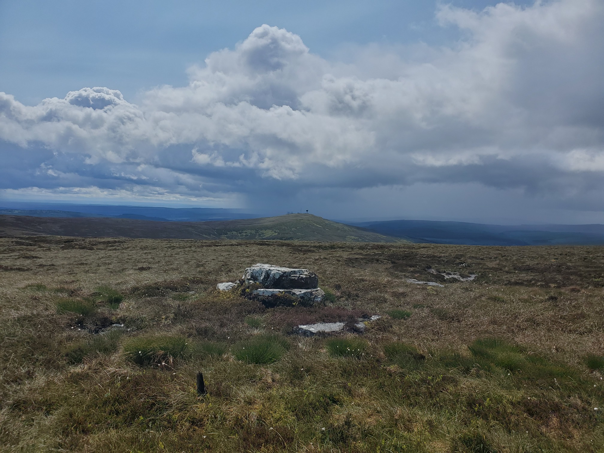 on top of the hill, a few stacked blocks of sandstone. The heather and bog extends into the distance, with the summit of Deadwater Fell behind, on it are some structures and a radar station. A very localise rainstorm is falling from clouds in the sky just behind Deadwater.