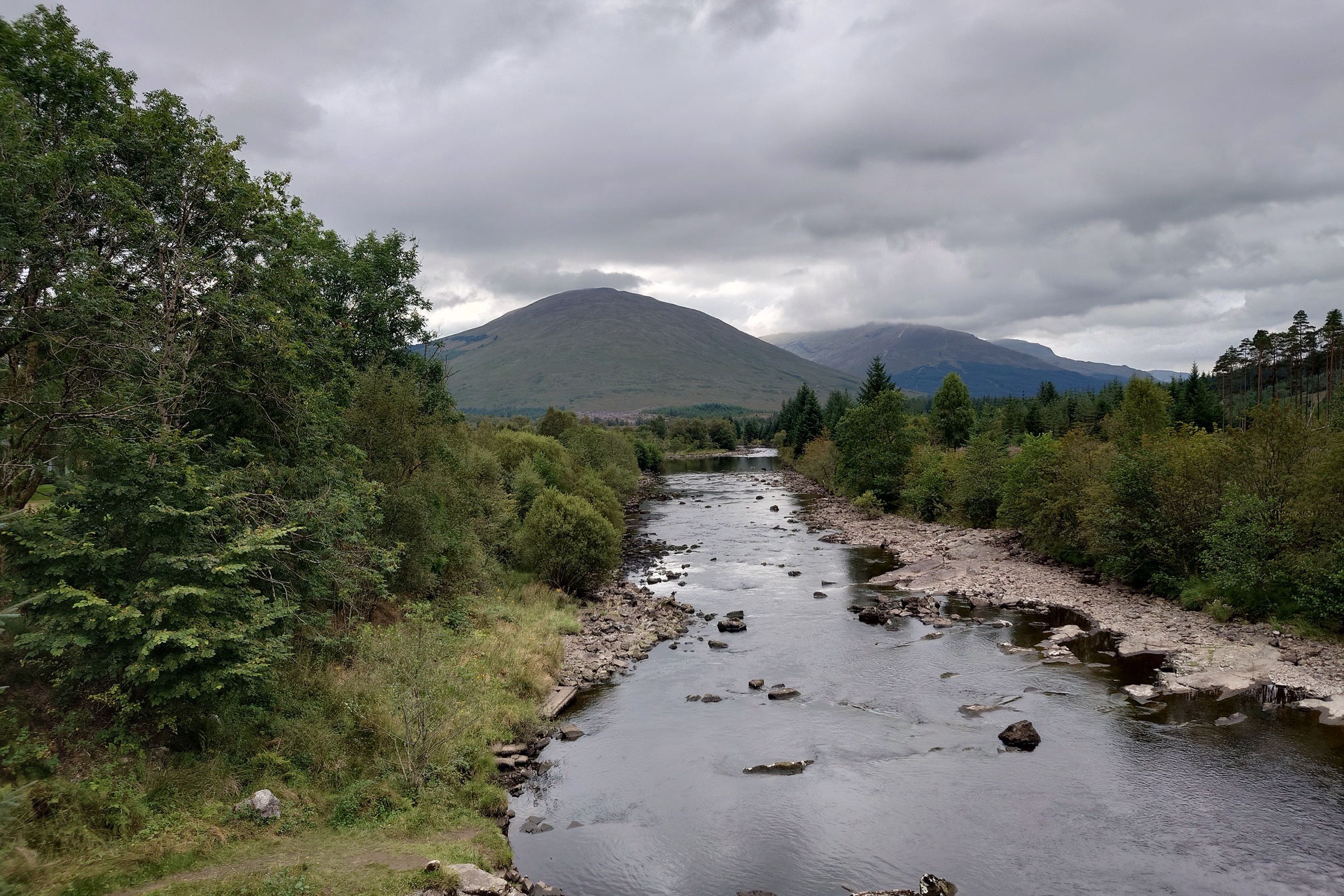 A river flows past, trees on both banks and mountains on the horizon