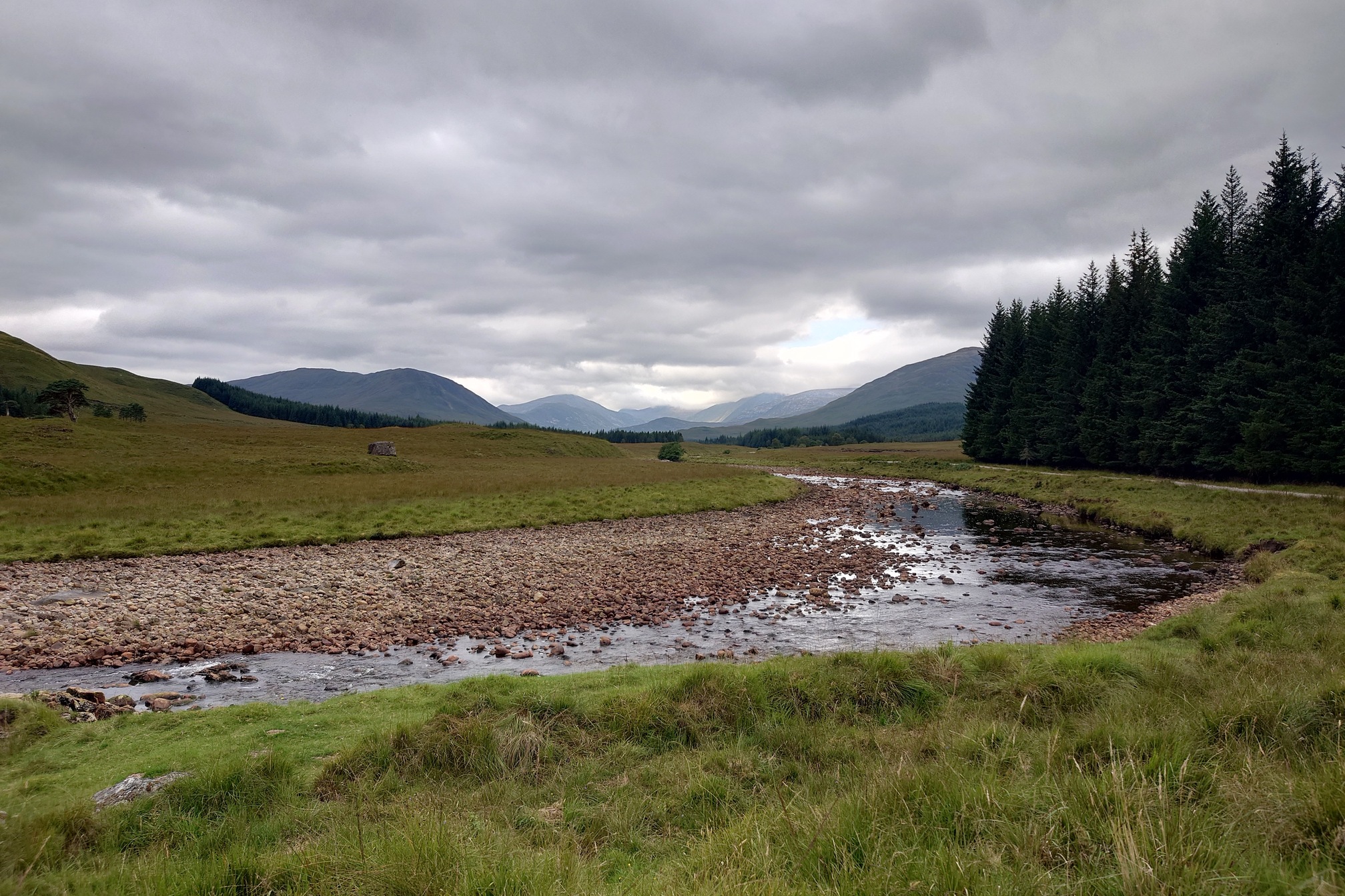 A broad, shalow rive flows over its stony bed. Grassland stertches away, through blocks of plantation forestry, towards numerous distant hills rising into the grey clouds