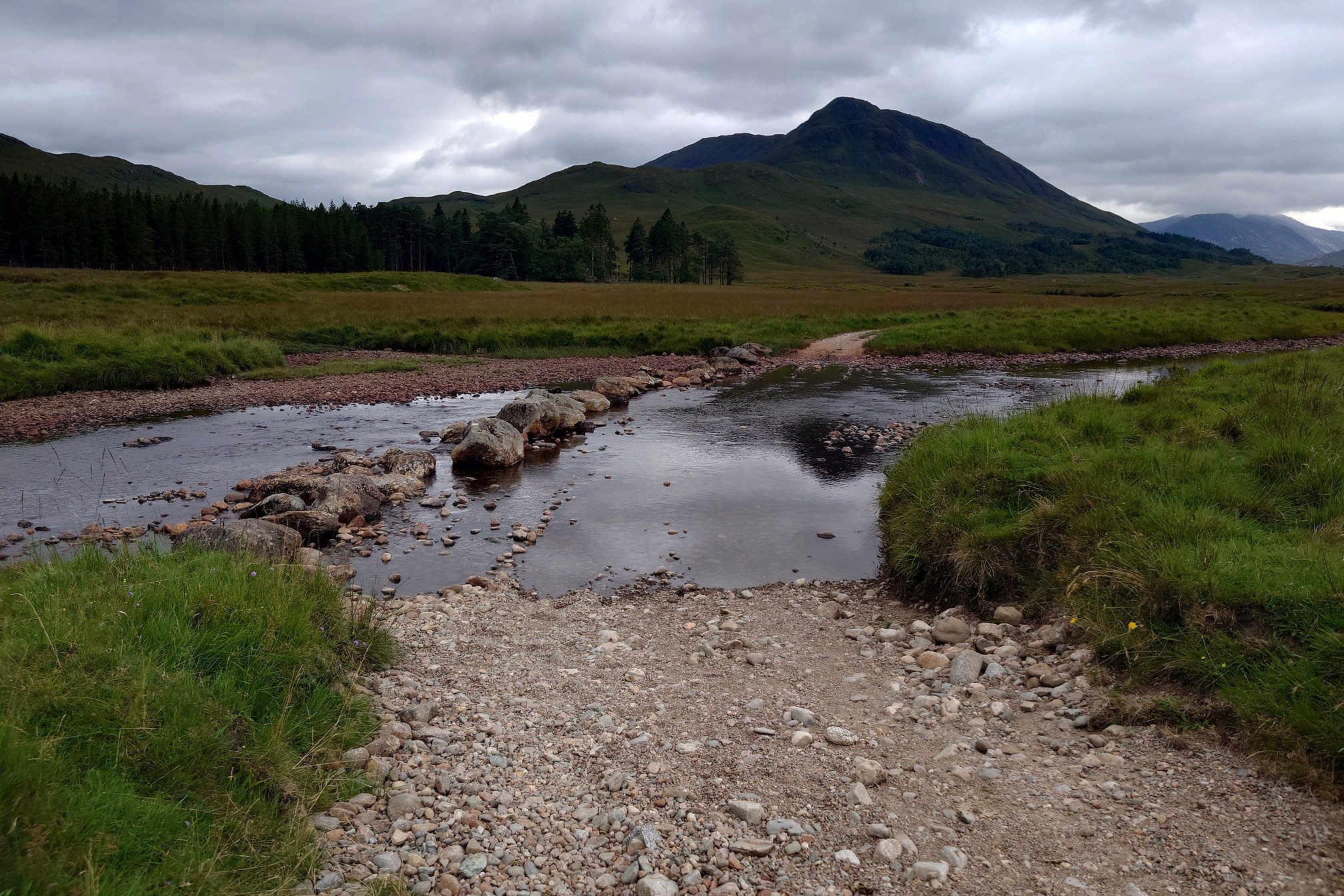 the gravel road leads straight into and across the river. Alongside it are a row of large rocks. The road curves to the right after crossing the river, with the large hill Beinn Sudhe behind.