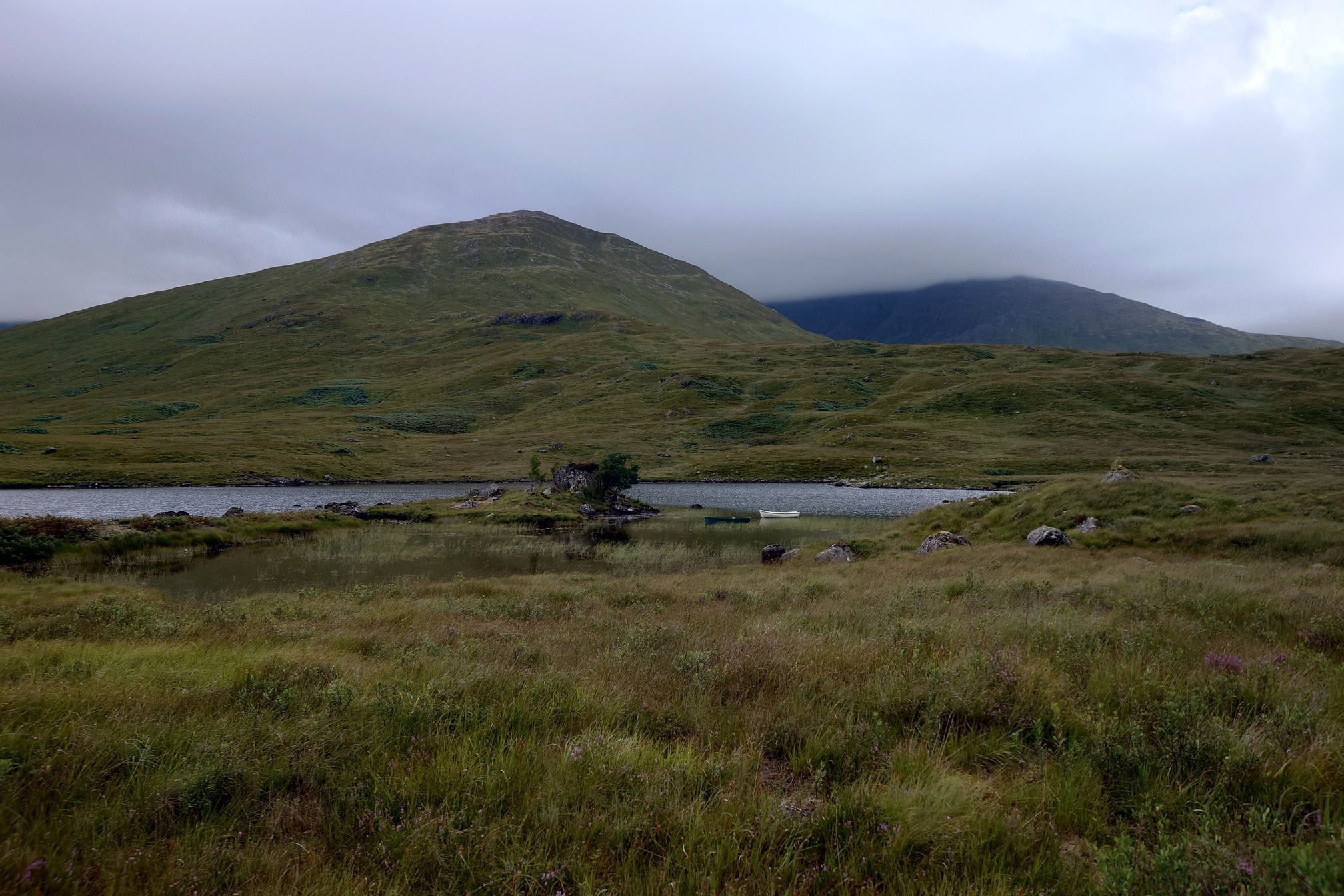 Grassy, rocky ground surrounds Loch Dochard. A hill rises up behind. A peninsula sticks out into the water, with a rocky outcrop and a couple of scrawny trees on it, and next to it are moored a green and a white wooden rowboat.