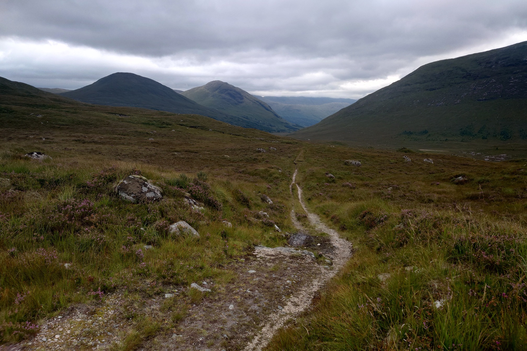 The track, now a narrow path, leads down through rocky and heathery ground into Glen Kinglass. Mountains either side.