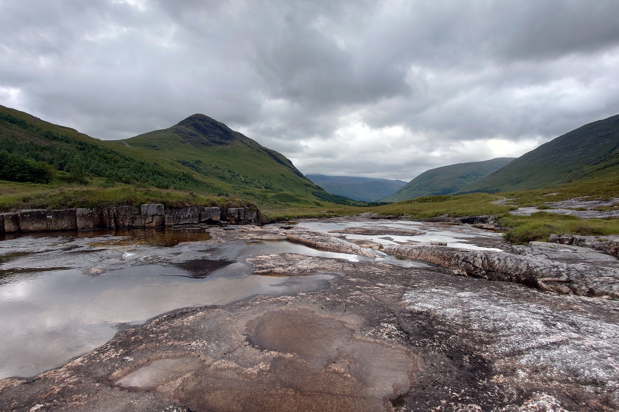 The river flows over flat rocks and outcrops between the mountains