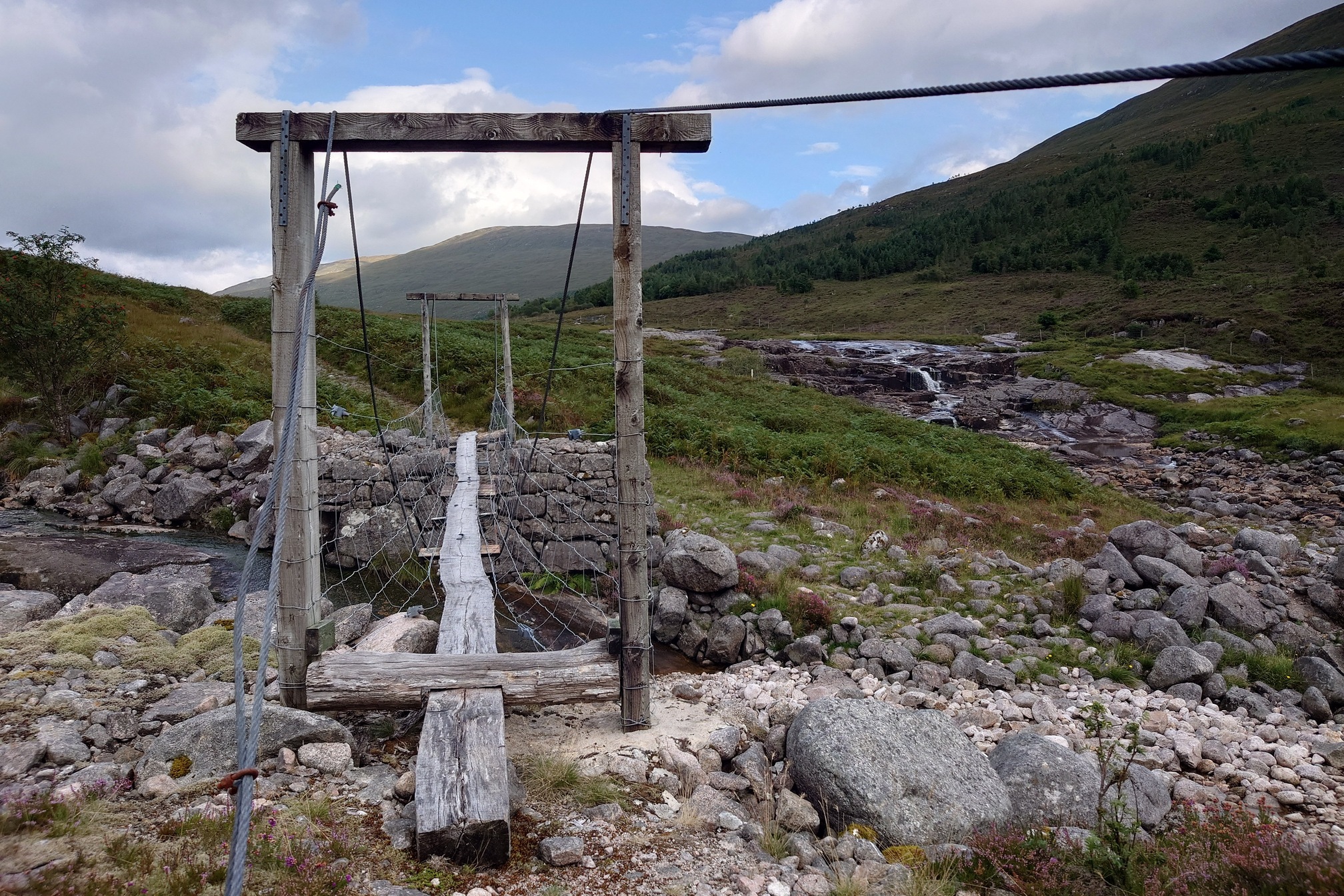 Looking across a very basic suspension footbridge. The bridge is a long, single beam of wood held up by cables from wooden uprights nailed together at either end. Sheep fencing strung along the side should stop you falling in. The ground is very rocky, and a water fall in the River Kinglass can be seen to the right of the picture. The stream the brdige crosses flows into this.