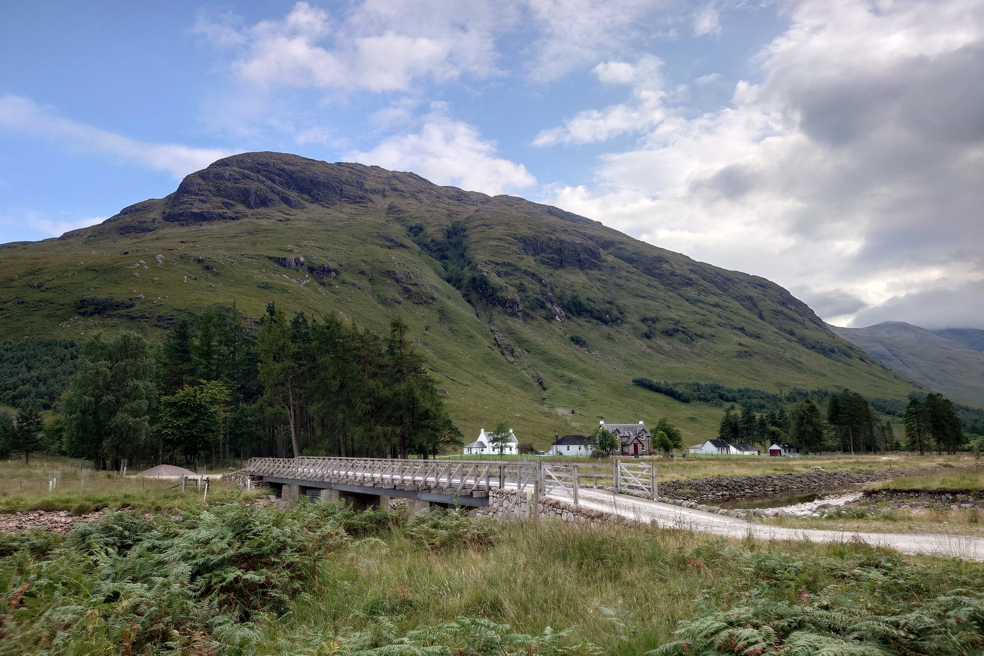 A small cluster of white-washed buildings and one larger stone house sit at the foot of Meal Garbh, a rocky and dramatic hill. Two patches of tall trees stand either side of teh buildings, and a road crosses the river on a substantial bridge to them