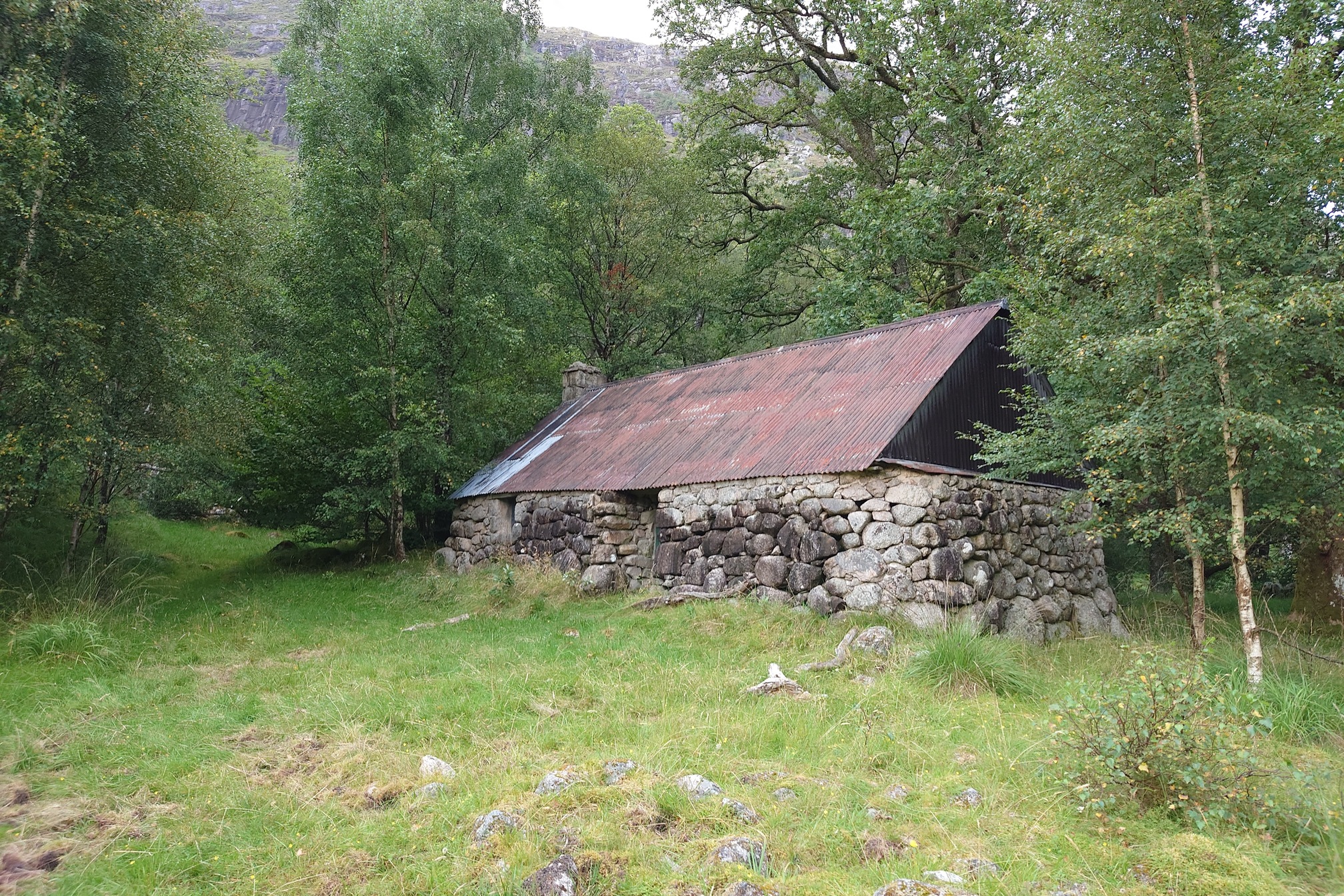A very old low stone building - a Black Hut - with a slightly more recent metal corrugated roof. It stands surrounded by trees and has a single window next to the door