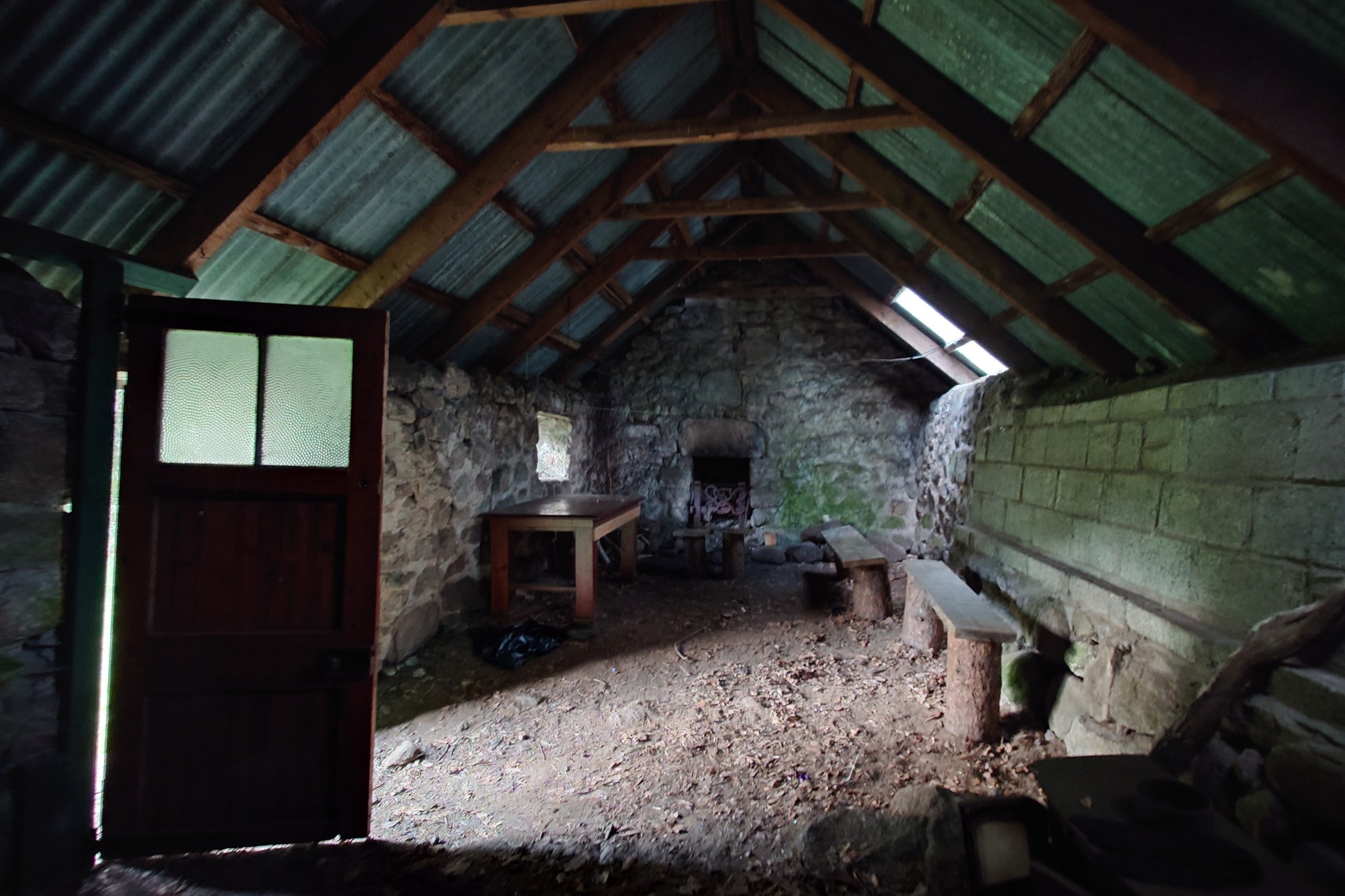 Inside, a fireplace in the end wall opposite, with dirt floor and a few crude stone benches, and a single table. A plastic skylight, the tiny window, and the open door are the only sources of light.