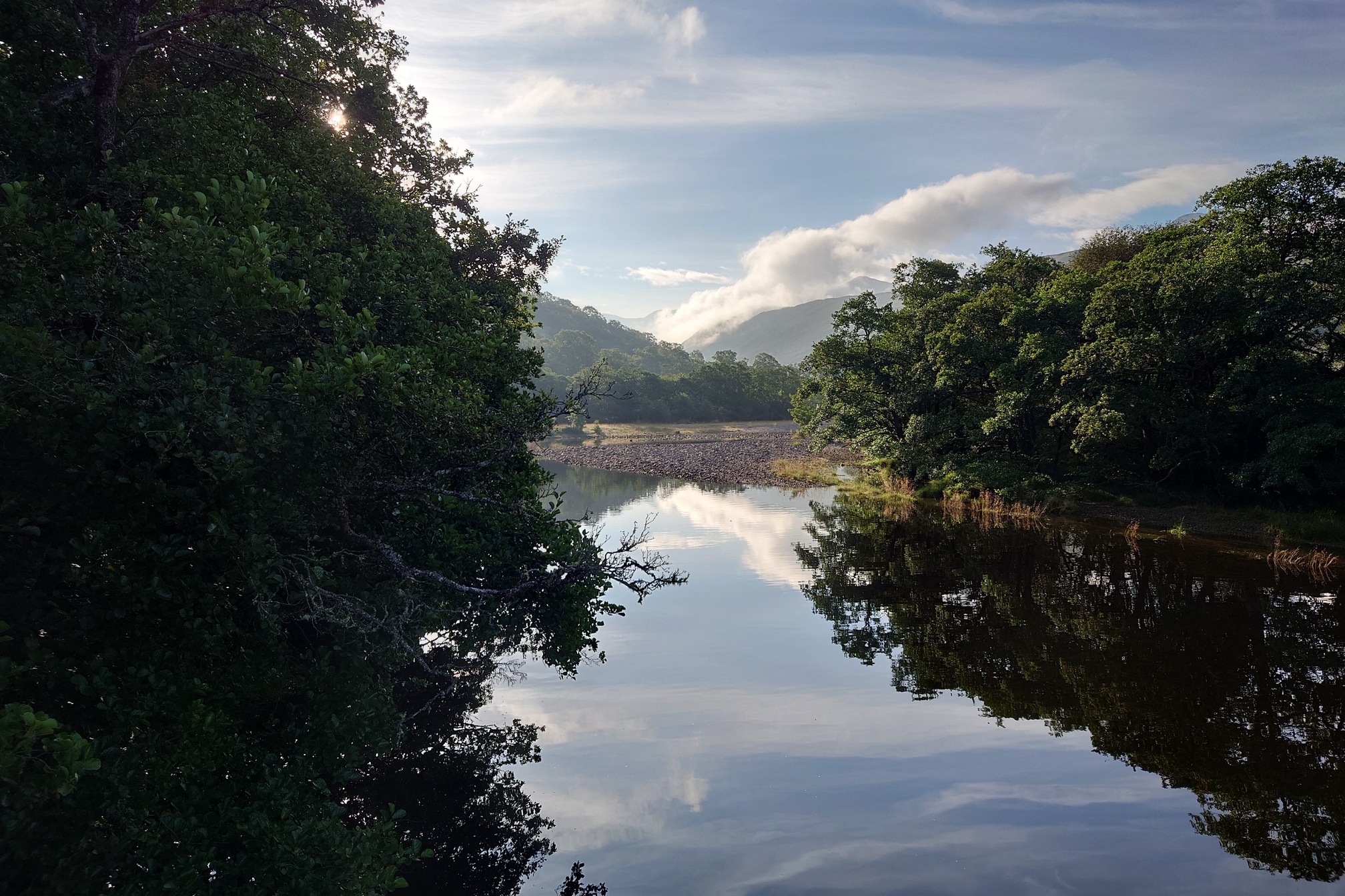 A broad, flat rive flows through trees towards a stony bar in the early morning light
