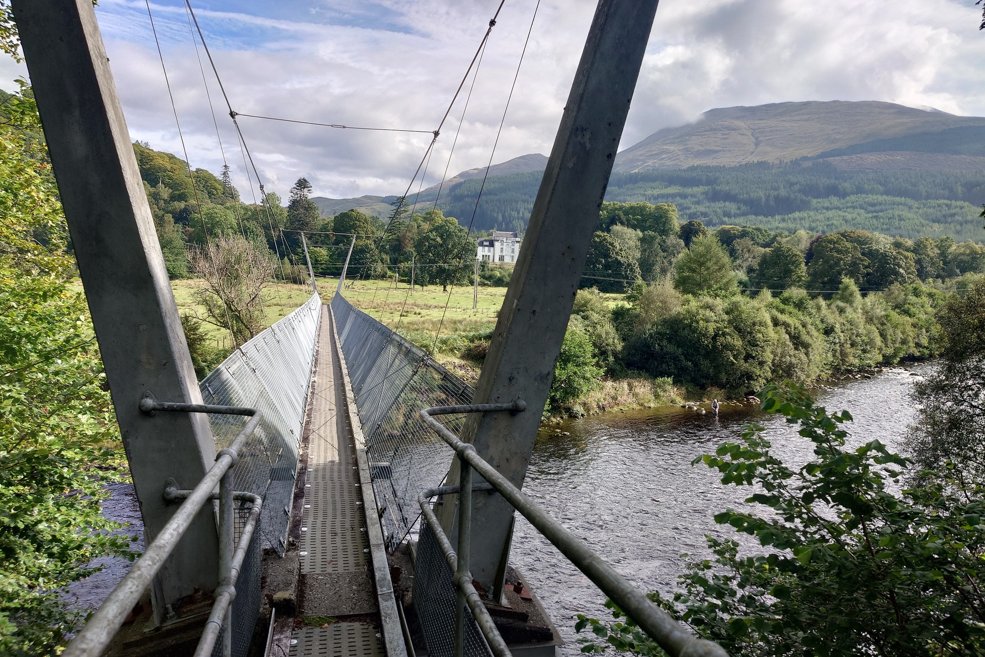 A narrow suspension footbridge crosses the river. In the woodland behind a white house, while stood in the river below is a flyfisherman.