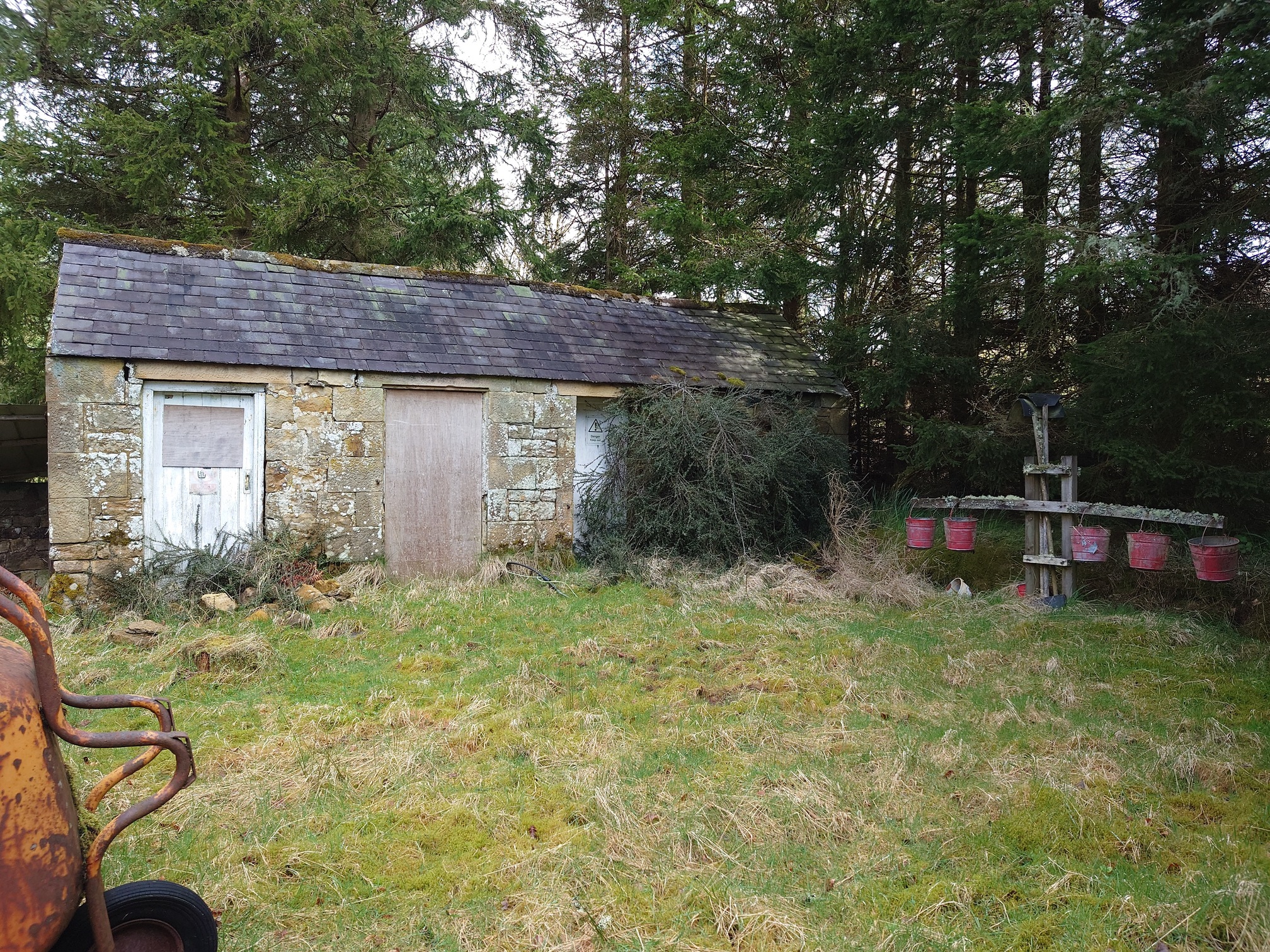 A low stone building with slate roof. The three doors we can see are boarded up, the ground in the foreground is very mossy and tussocky. To the right is a wooden cross structure: in teh centre are rotten old firebeaters, while hanging from the arms either side are five red fire buckets. Behind are very tall trees.