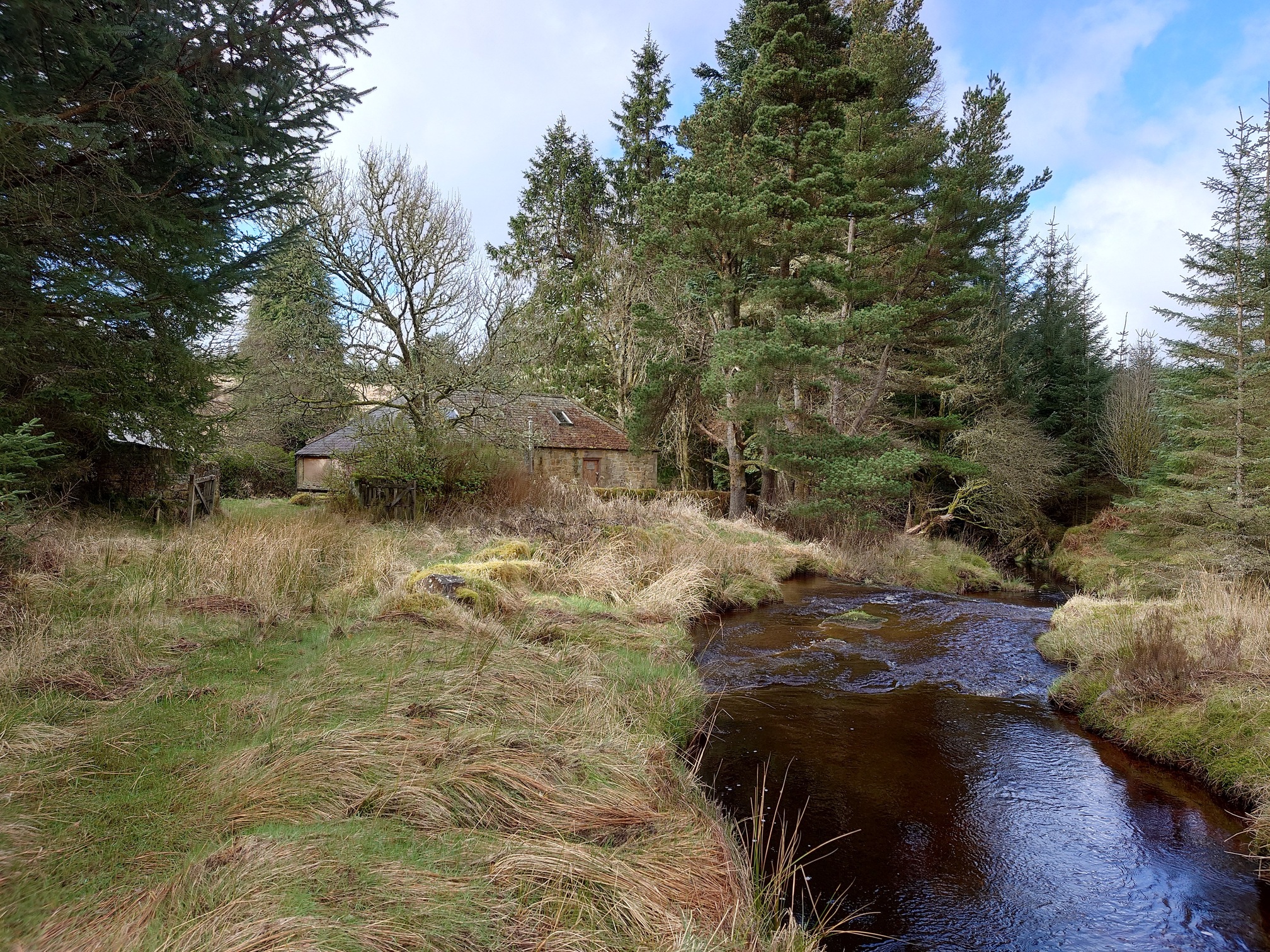 the burn flows to the right of the image, on the left, surrounded by tall trees, a low stone building with a metal chimney sits behind a wall. Nearby, a gate is open and half off its hinges