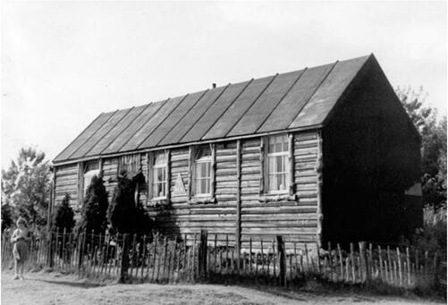 A black and white photo of a neat, simple wooden building. It has four windows and a door partly obscured by ornamental bushes. A low wooden fence surrounds it, and a young woman is stood on the far left of the picture.
