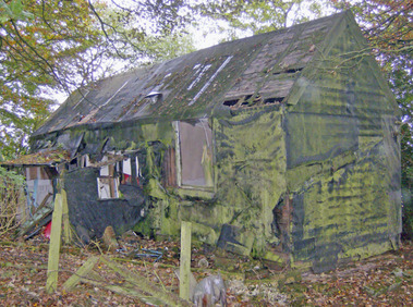 The same building, but a more modern photo (2005). It is extremely ramshackle, the roof has holes, and the whole exterior at some point has been covered with black material, possibly tarpaulin, which is now covered in moss and falling down. A porch was added at some point, and this is now also falling down. The entire site is surrounded by trees.