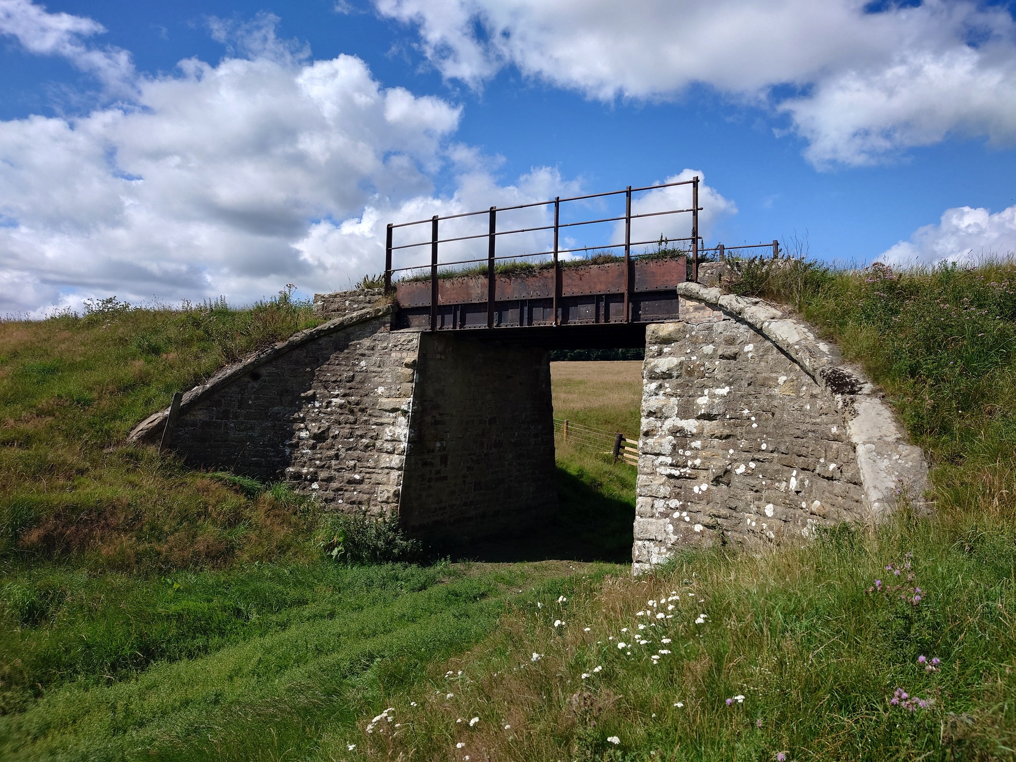 another railway bridge crosses a farm track. The stone supports either side curve gracefully round, and the trackbed is carried on metal girders over the path.