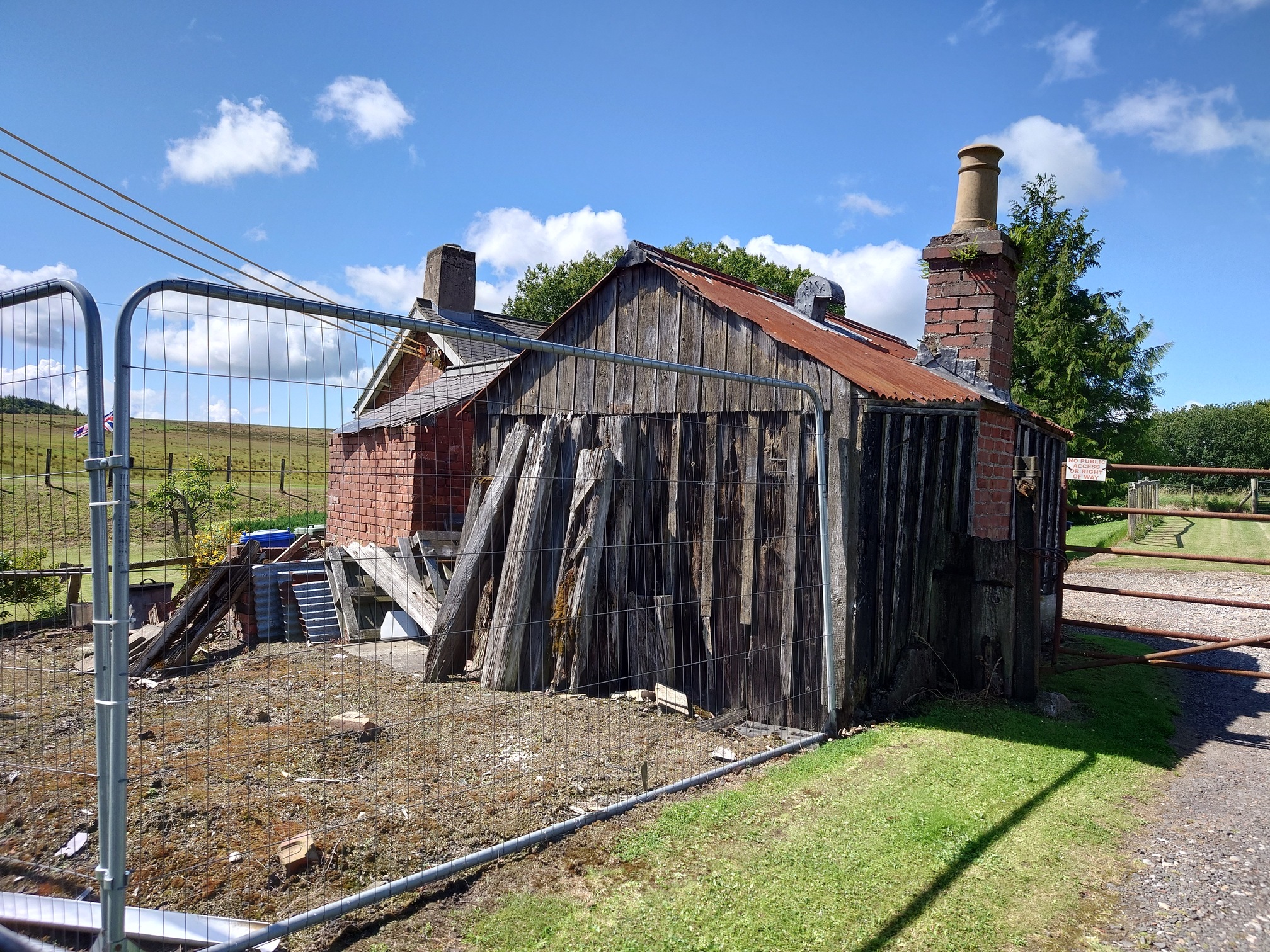 a very old and rotten railway hut, made of upright wood with a rusty corregated metal roof and a wonky brick chimney. It's safely fenced off with security fence and it looks like someone is working on maintaining it. Behind can be seen more brick buildings.