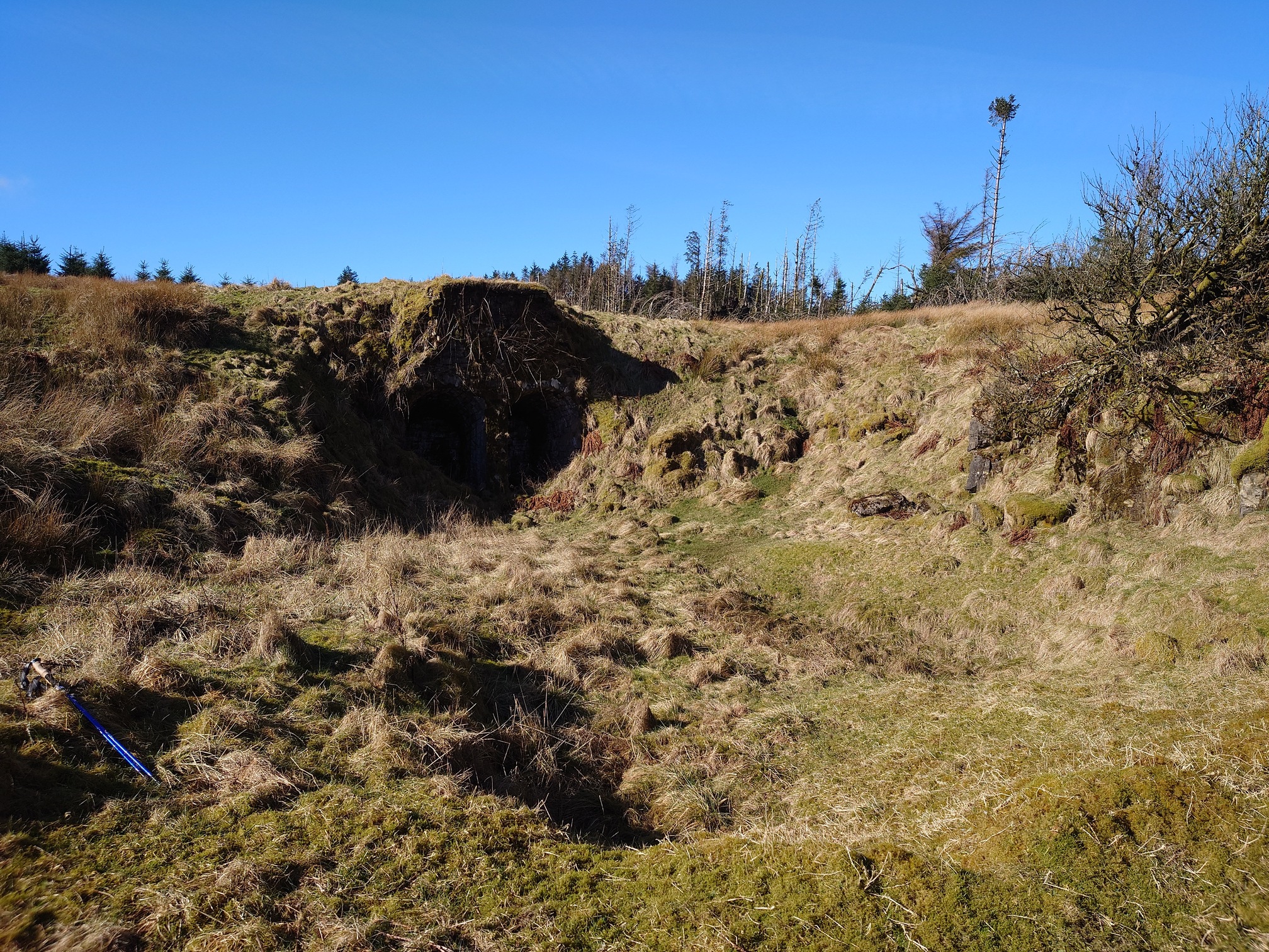 At the bottom of a grassed-over quarry. Across the uneven ground, hidden in shadow as the sun is out now, can just be made out two large arches leading into the quarry wall. l
