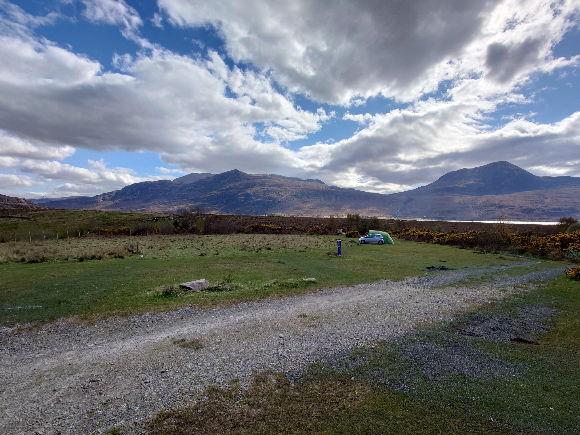 a tiny car and a tent a little bit bigger behind it are pitched on a flat grass campsite, bordered by flowering gorse. Behind mountains rise up